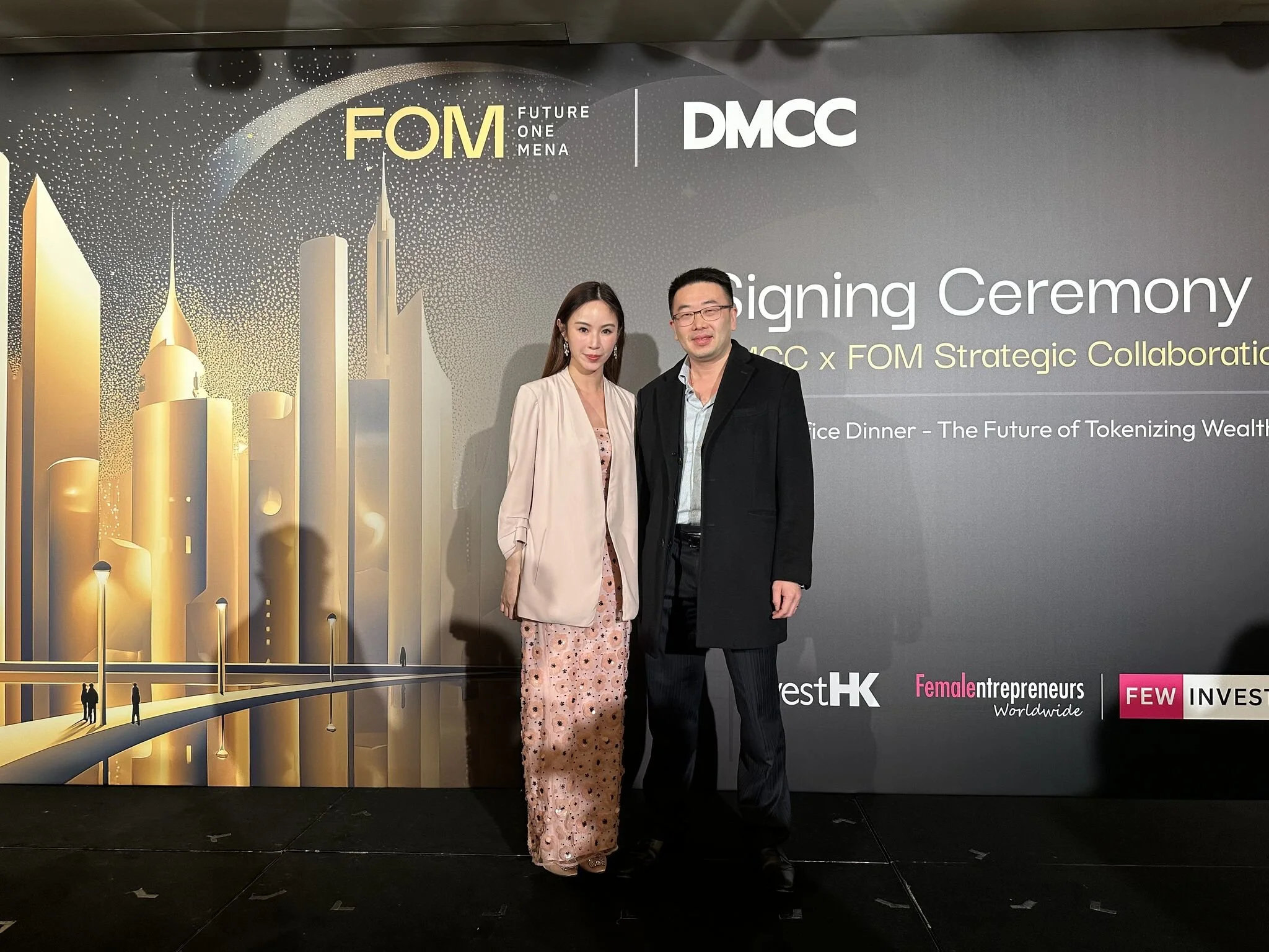 Two people, a woman and a man, standing on a stage in front of a backdrop at a signing ceremony event. The backdrop features logos for FOM and DMCC, and text indicating a strategic collaboration. The woman is wearing a pink floral dress with a beige blazer, and the man is in a dark suit with a light shirt. The stage has a black floor, and the background displays a futuristic cityscape illustration with tall golden buildings and lights.