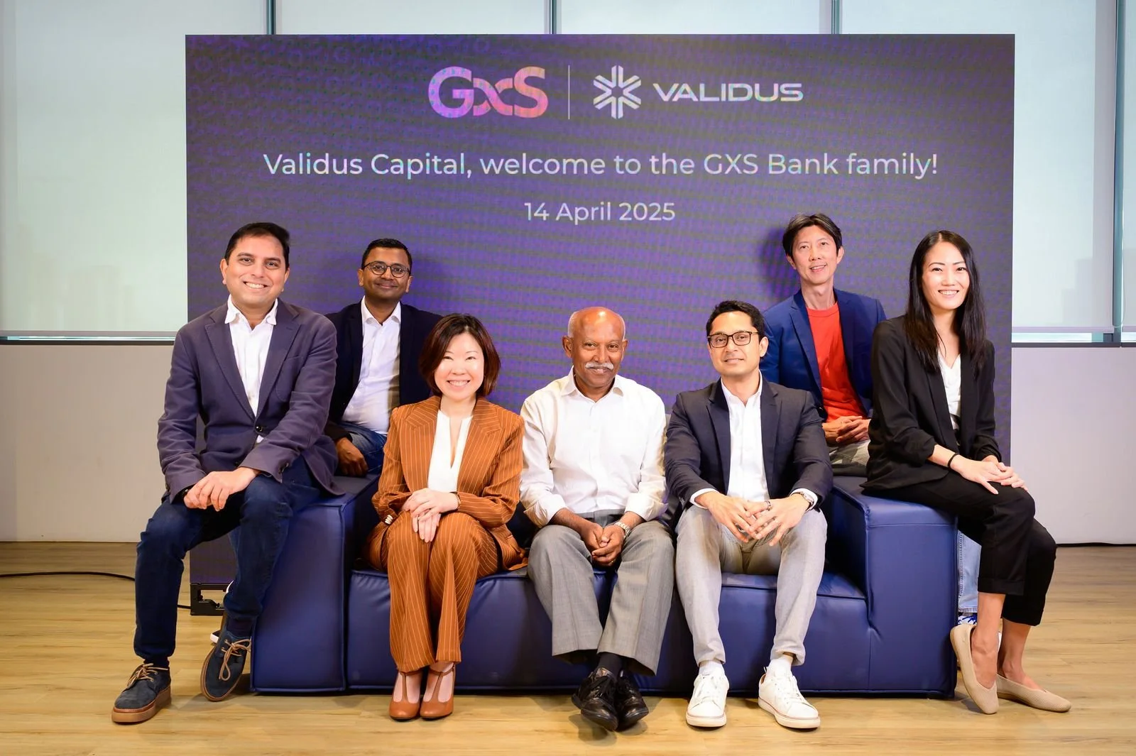 Group of seven diverse professionals sitting and standing in front of a digital screen displaying the logos of GXS and Validus, with the message "Validus Capital, welcome to the GXS Bank family! 14 April 2025" in a modern office setting.