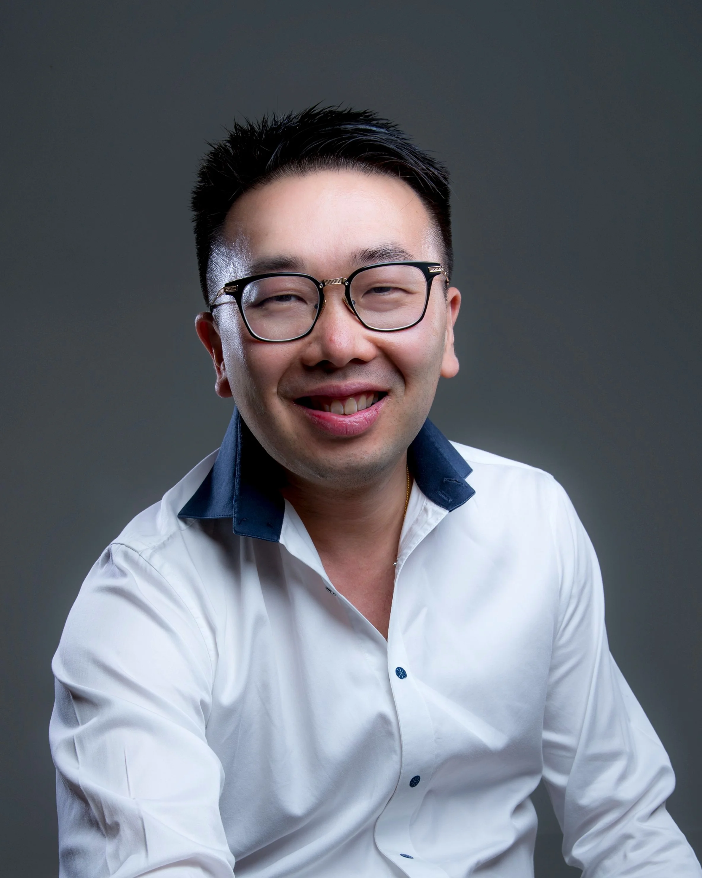 A smiling man with glasses wearing a white shirt with dark collar detail, posing against a plain dark background.