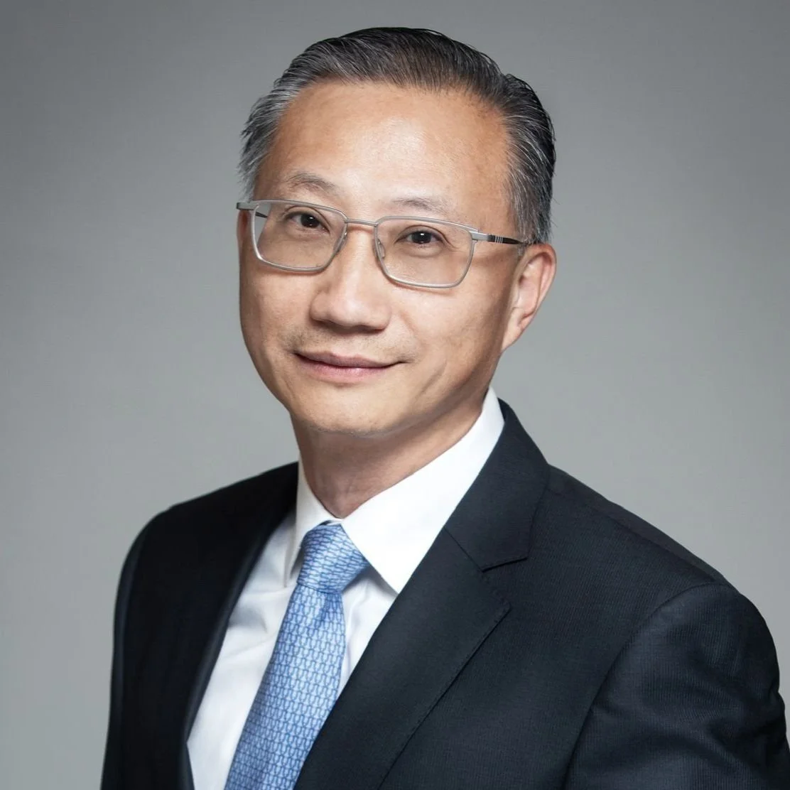 Professional portrait of a middle-aged Asian man wearing glasses, a dark suit, white shirt, and blue tie, smiling against a gray background.