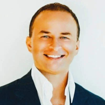 Close-up photo of a smiling man with short dark hair wearing a white shirt and dark blazer.
