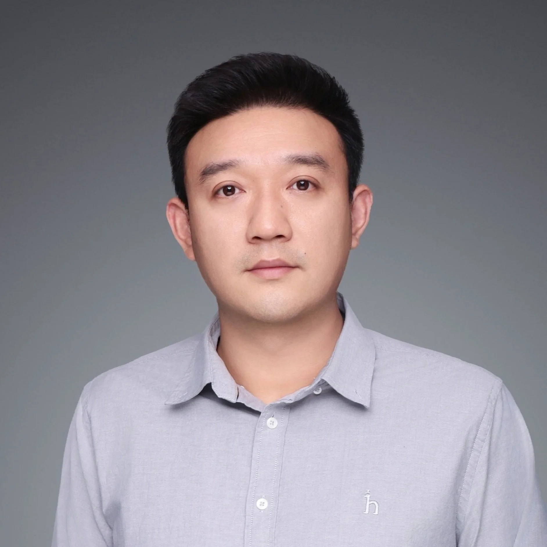 A portrait of a young Asian man with short black hair wearing a light gray button-up shirt against a gray background.