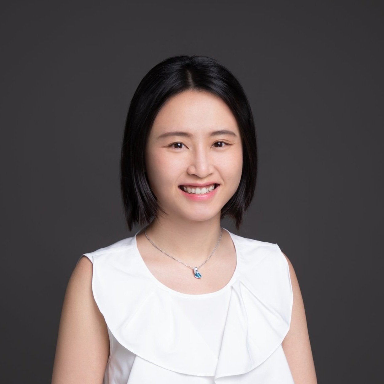 A portrait of a young woman with short black hair, smiling, wearing a white sleeveless top and a silver necklace with a blue pendant, against a dark grey background.