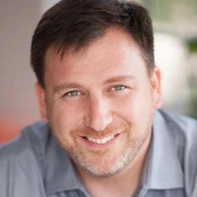 Close-up of a smiling man with short brown hair and a beard, wearing a gray shirt.