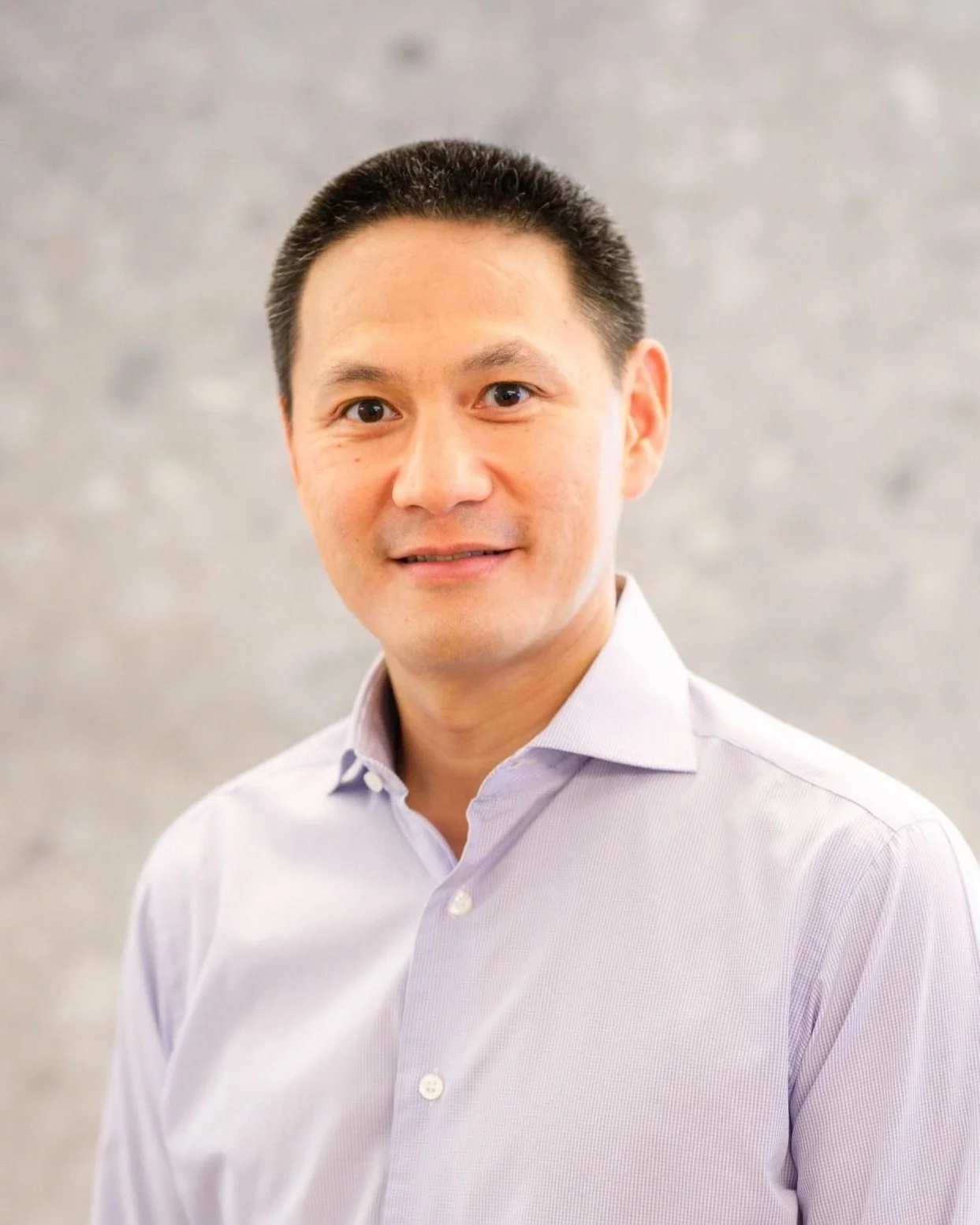 A portrait of an Asian man in a light purple button-up shirt, smiling against a blurred grey background.