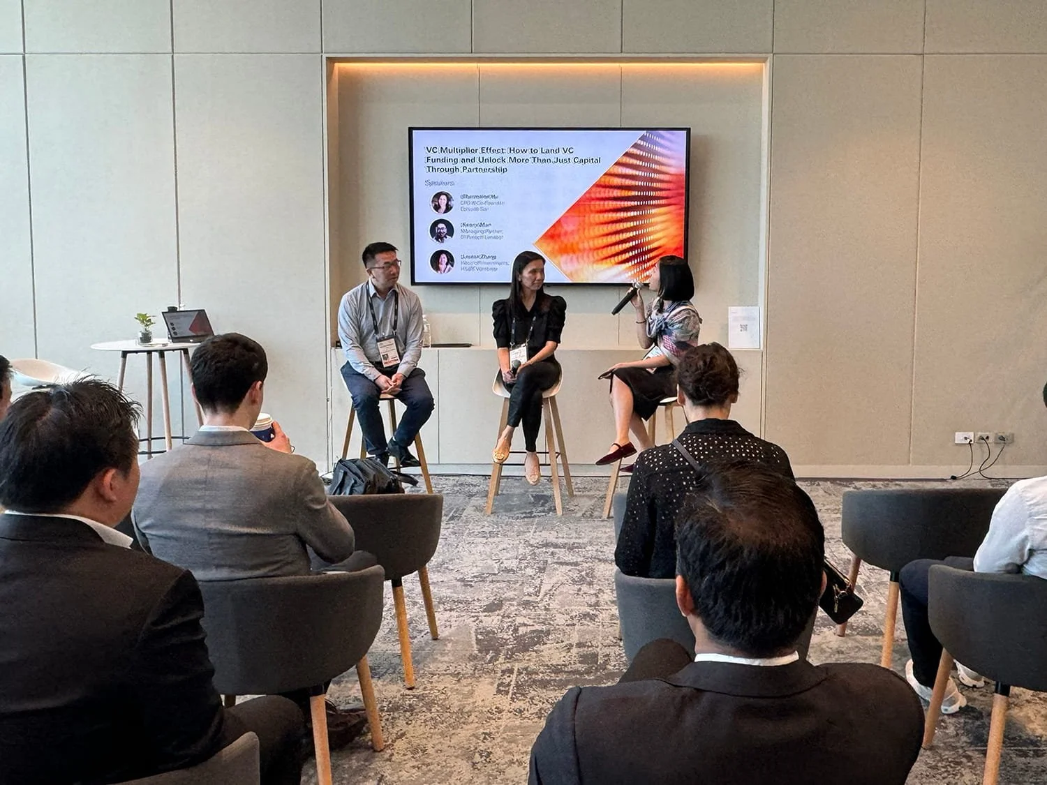 A panel discussion at a conference with three women seated on high chairs on stage, one woman speaking into a microphone, with an audience seated in front. A large screen behind them displays a presentation titled 'VC Multiplier Effect: How to Land V