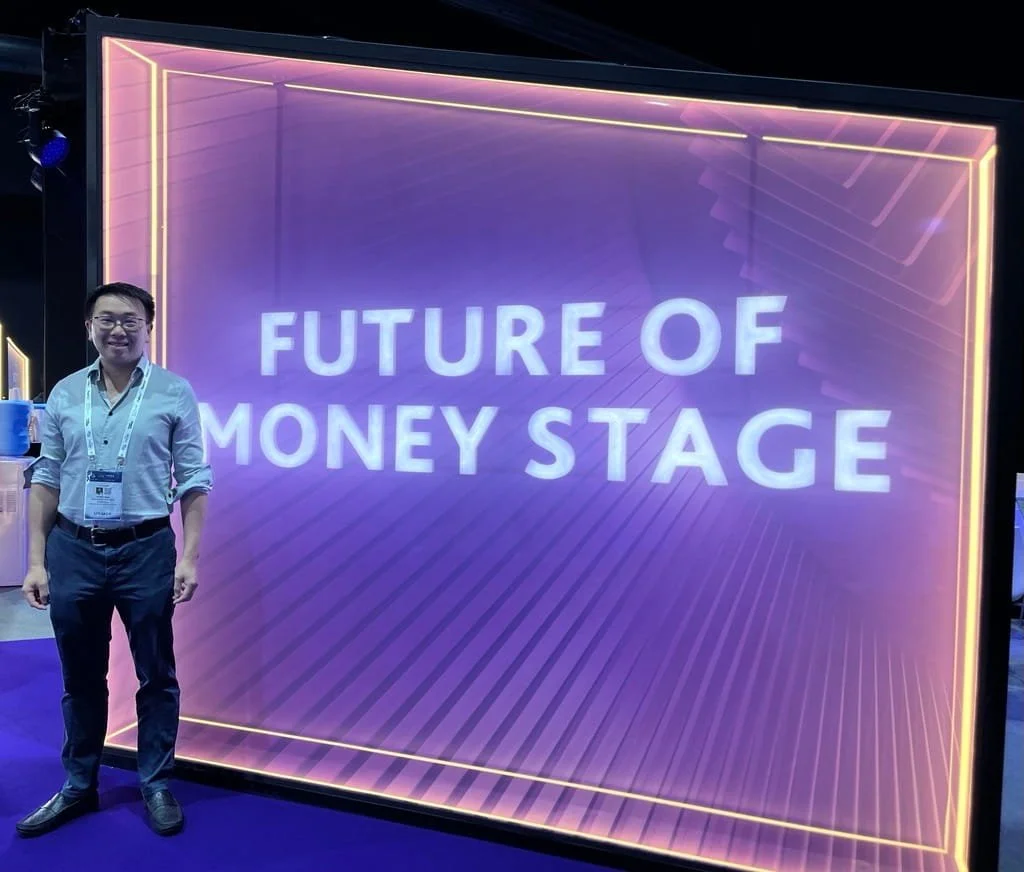 A man wearing glasses and a white shirt stands next to a large, illuminated sign that reads "FUTURE OF MONEY STAGE" at a conference or event.