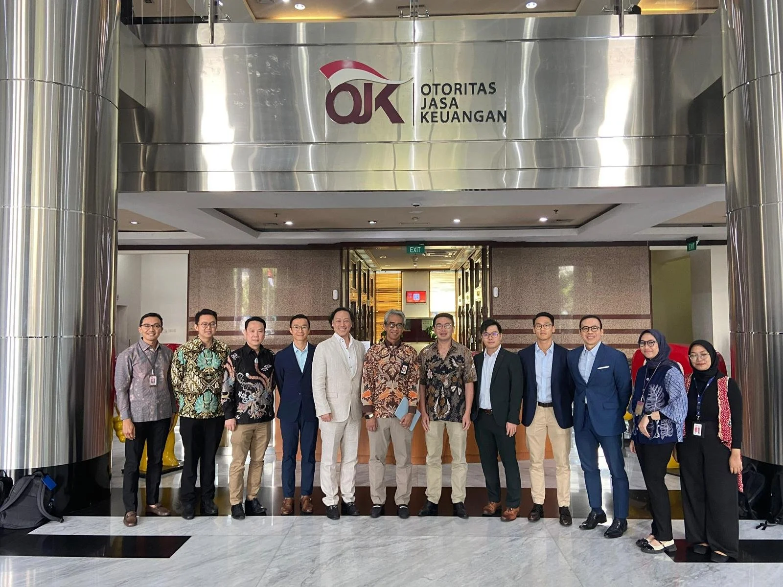 Group of people standing in a line in front of the entrance to OJK (Otoritas Jasa Keuangan) building, dressed in business attire, with some wearing traditional batik shirts.