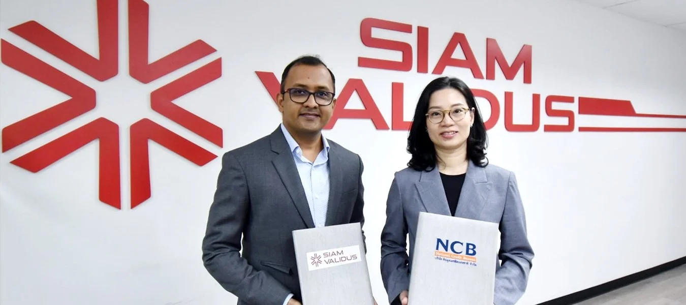 Two business professionals, a man and a woman, standing in front of a wall with the 'SIAM VALIDUS' logo and a geometric snowflake design. They are holding documents with the logos 'SIAM VALIDUS' and 'NCB'.