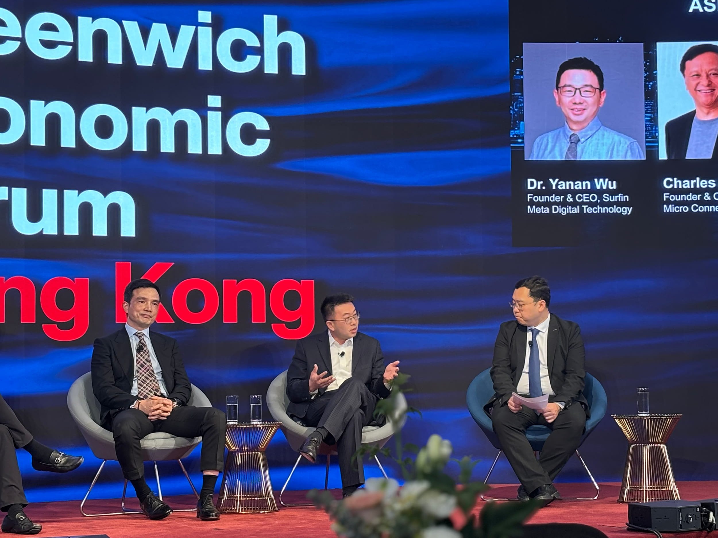 Three men are seated on stage during a panel discussion at a conference about Hong Kong's economy. The background screen displays the words 'Hong Kong Economic Forum' with photos and names of two speakers, Dr. Yanan Wu and Charles. The man in the mid