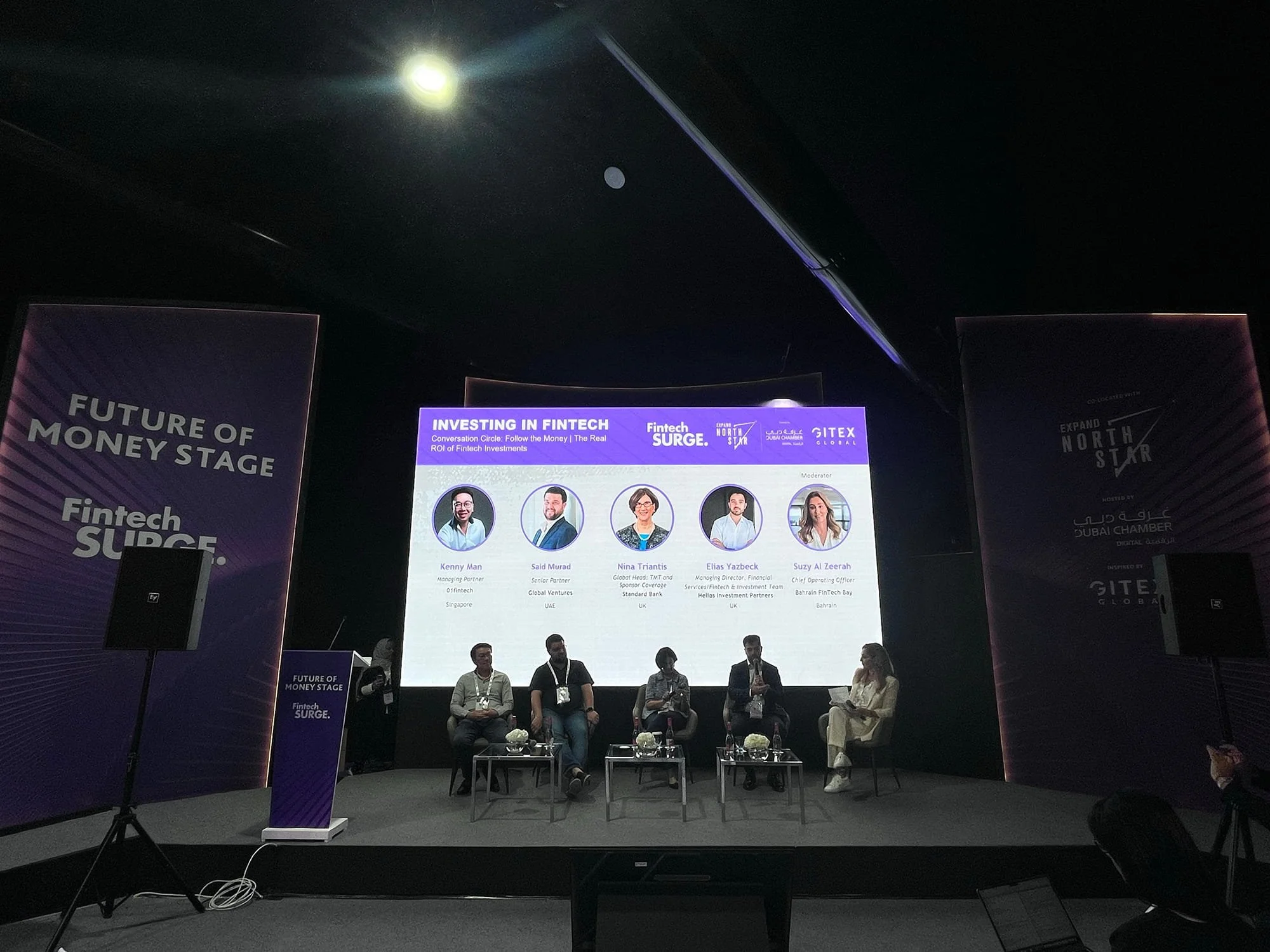 Panel discussion at a conference stage with five speakers seated in chairs, a large digital screen behind them displaying information about the event and speakers, and banners on the sides with text about the event, titled 'Future of Money Stage' and