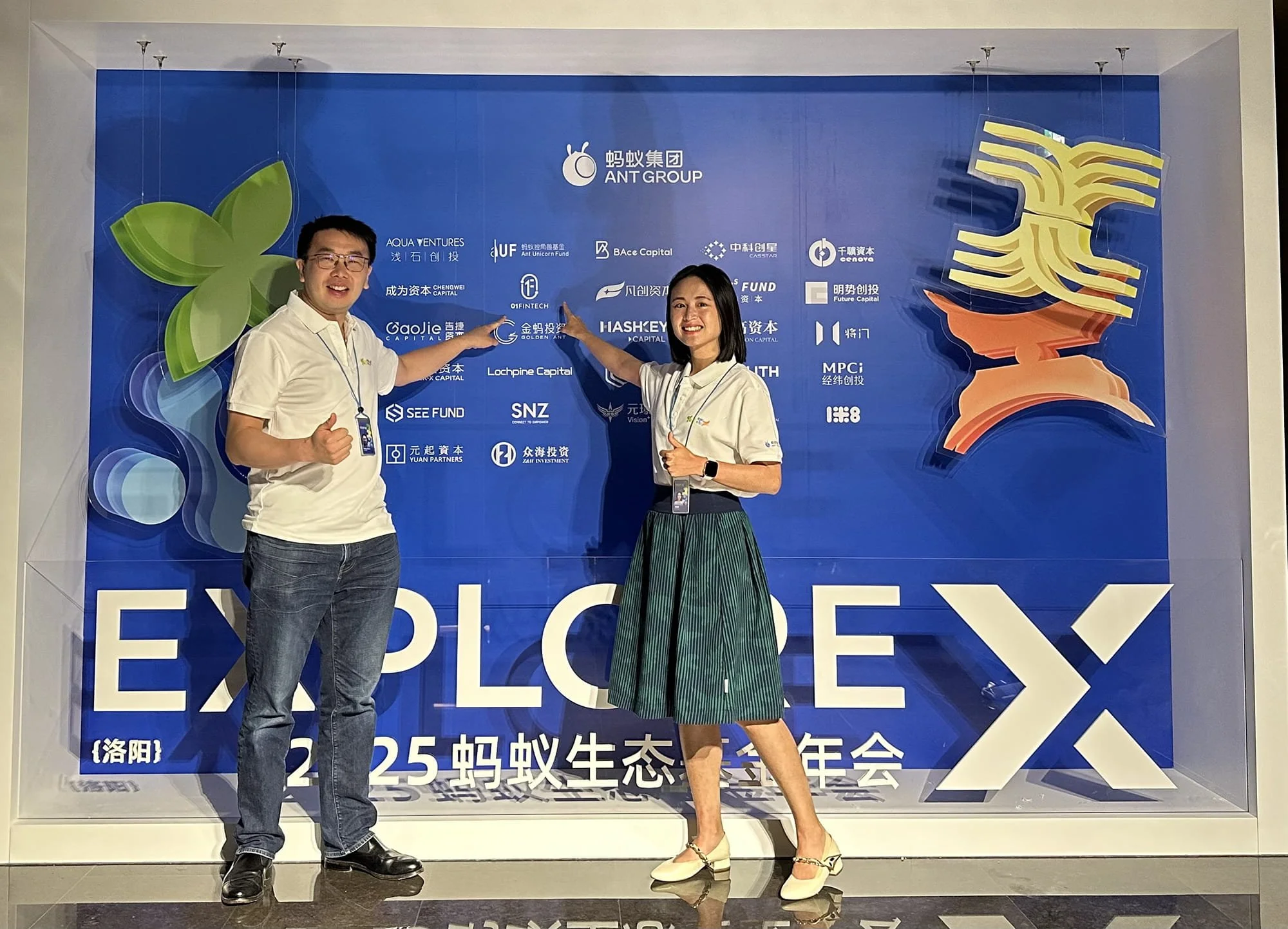 Two people posing in front of a blue event backdrop with logos and Chinese characters. The man on the left is giving a thumbs-up, wearing glasses, a white polo shirt, jeans, and black shoes. The woman on the right is smiling, wearing a white polo shirt, a green striped skirt, and beige heels, pointing toward the backdrop.