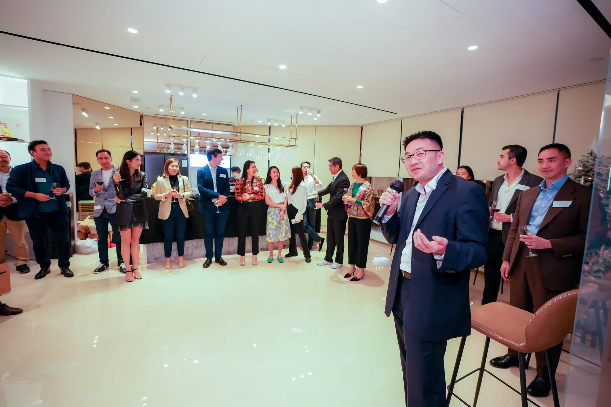 A man holding a microphone and speaking to a group of people at a social event or cocktail party in an indoor venue with warm lighting.