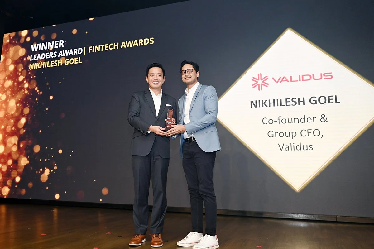 Two men in suits stand on stage, holding an award together, smiling. Behind them, a large screen displays that Nikhilesh Goel is the winner of the Leaders Award in the Fintech Awards, representing Validus.