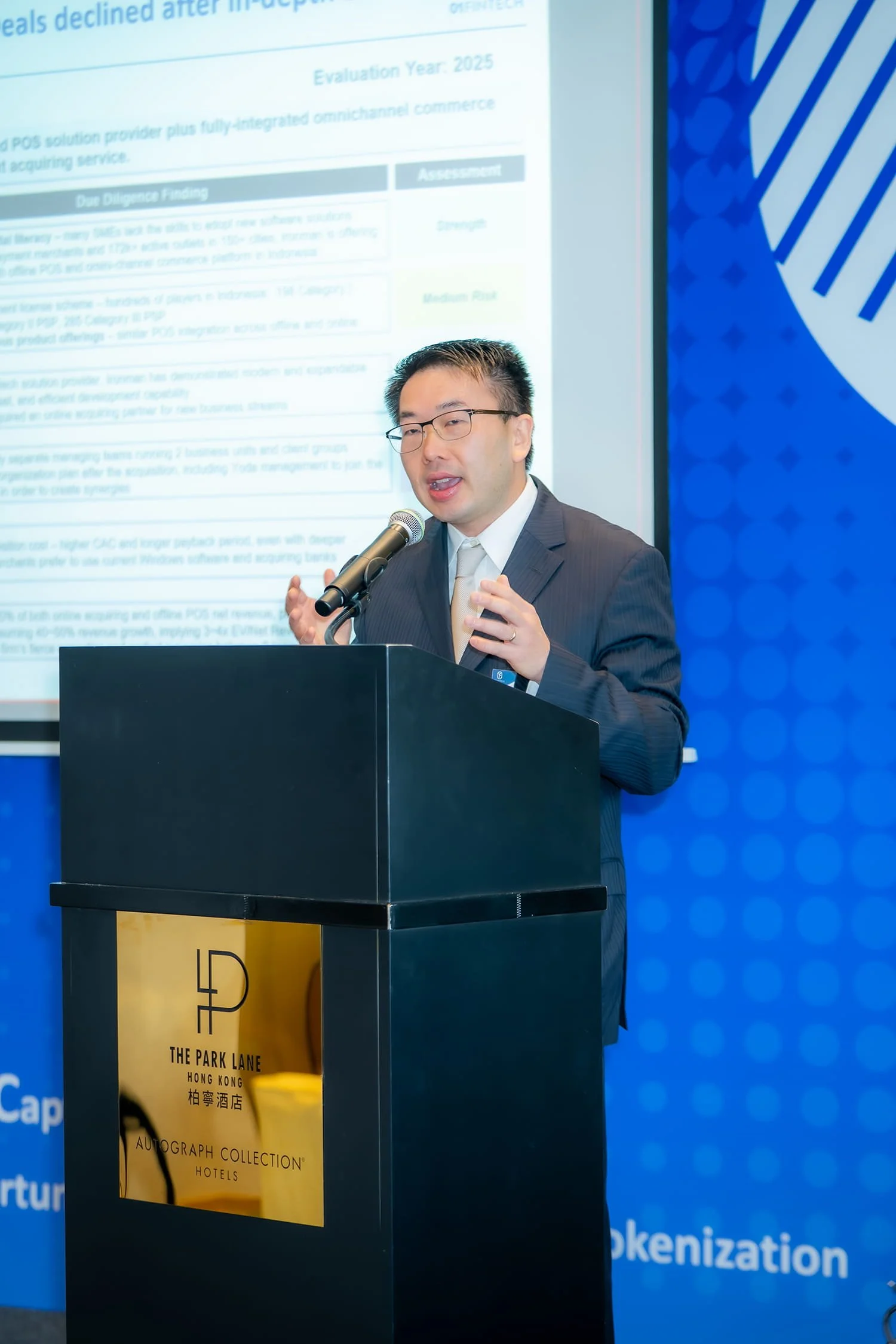 A man in a dark suit and tie speaking at a podium with a microphone, with a large projected presentation slide behind him. The slide contains text and assessment details, and the podium has a logo for The Park Lane Hong Kong Autograph Collection Hote
