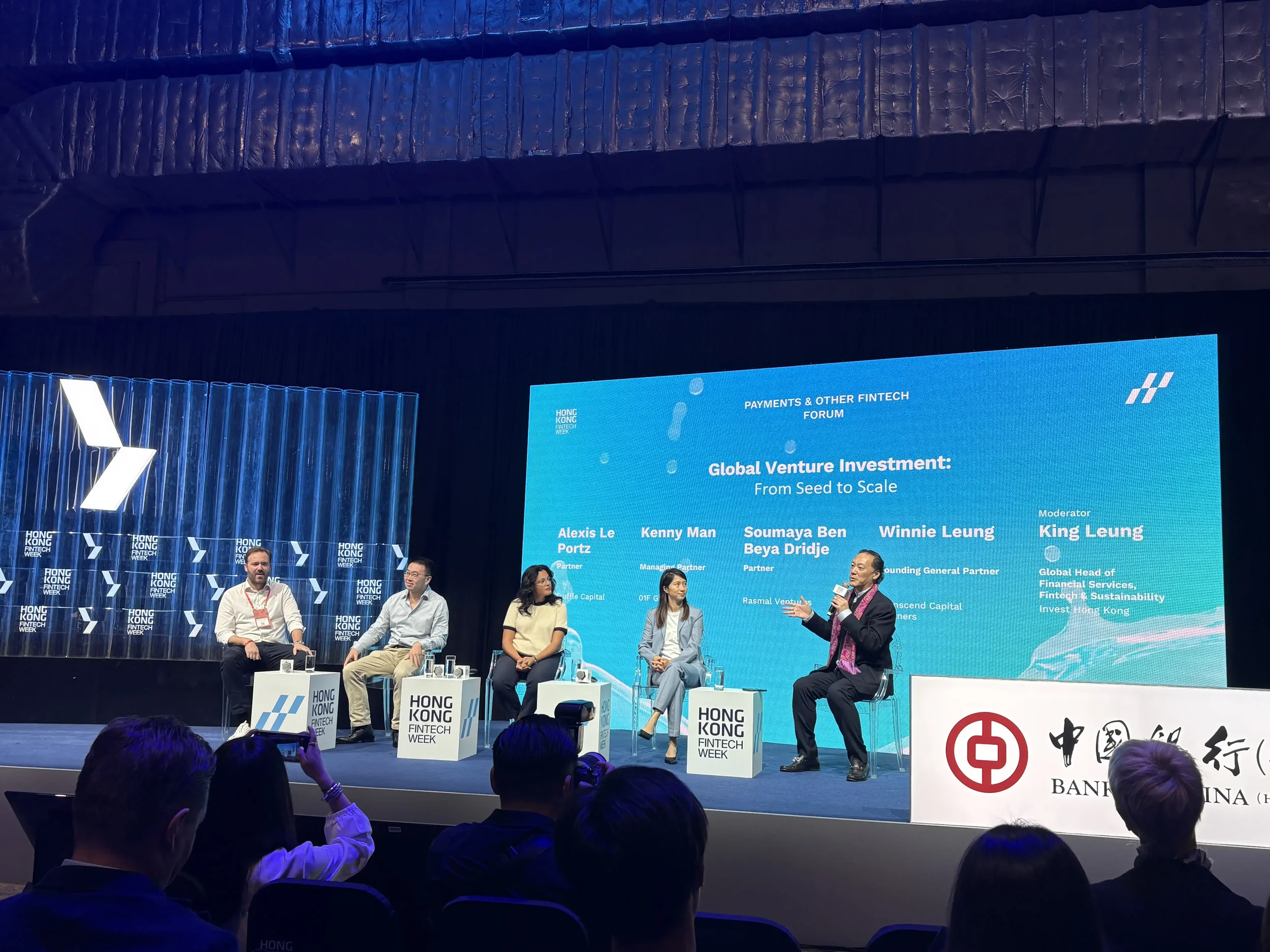 Panel discussion at Hong Kong Fintech Week with five speakers on stage, five panelists seated in front of a large blue screen displaying event information and speaker names, audience watching in the foreground.
