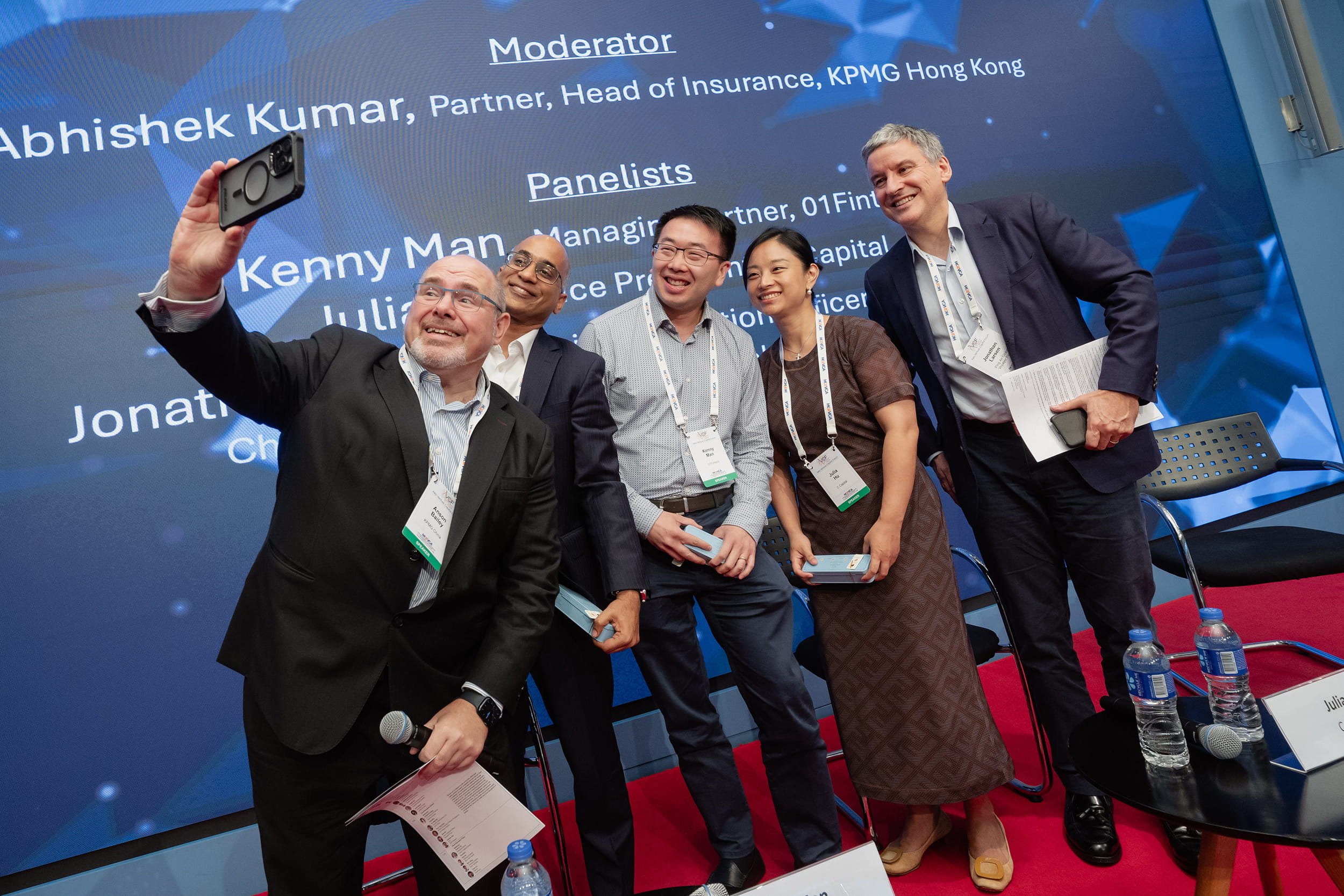 Group of five diverse professionals, including four men and one woman, taking a selfie together at a conference or panel event. They are smiling and holding notebooks or papers, standing in front of a large screen displaying names and titles of the m
