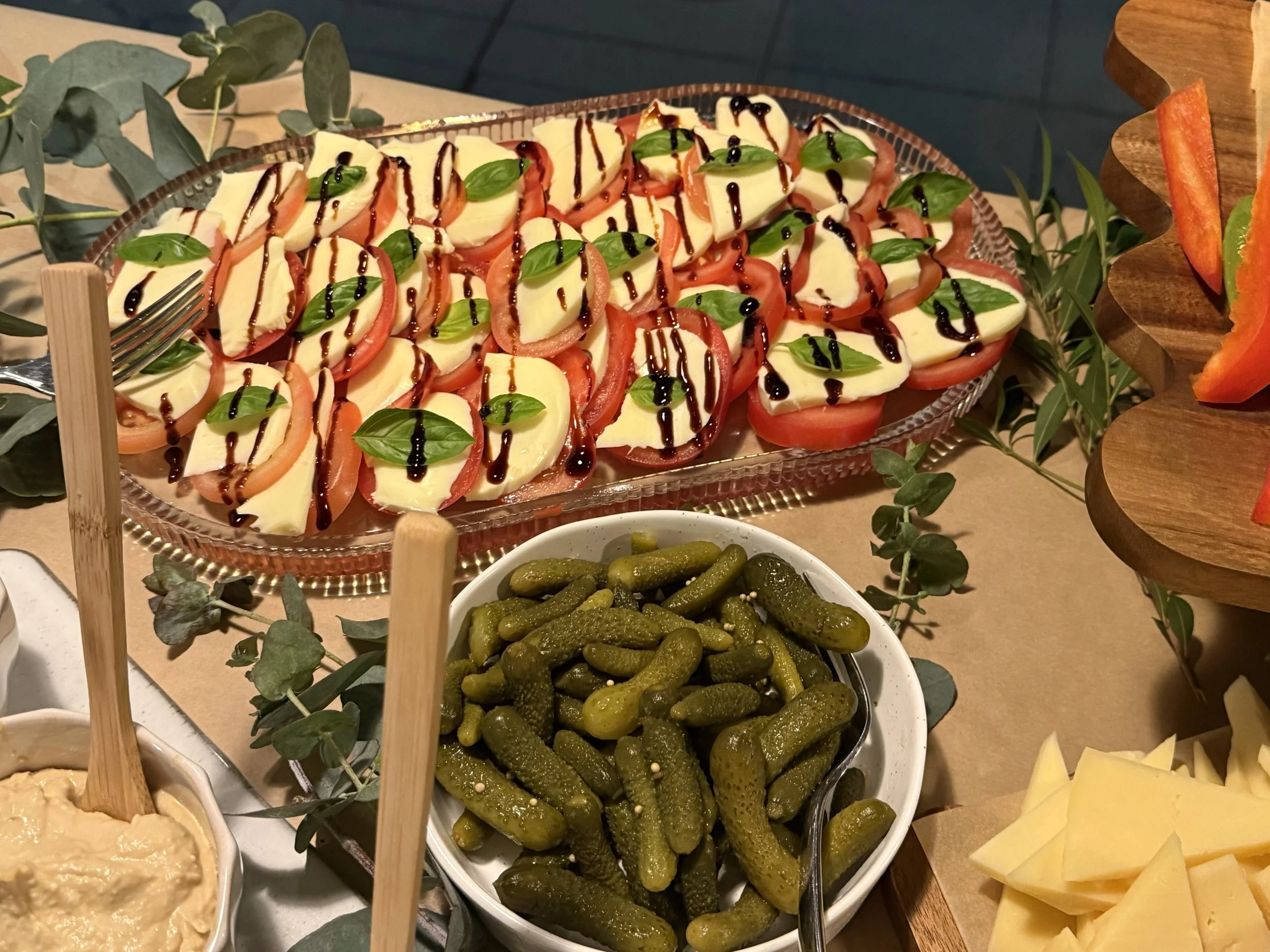 A plate of sliced mozzarella cheese and tomato slices garnished with fresh basil leaves and drizzled with balsamic glaze. Surrounding bowls contain pickles, a creamy dip, and slices of cheese.