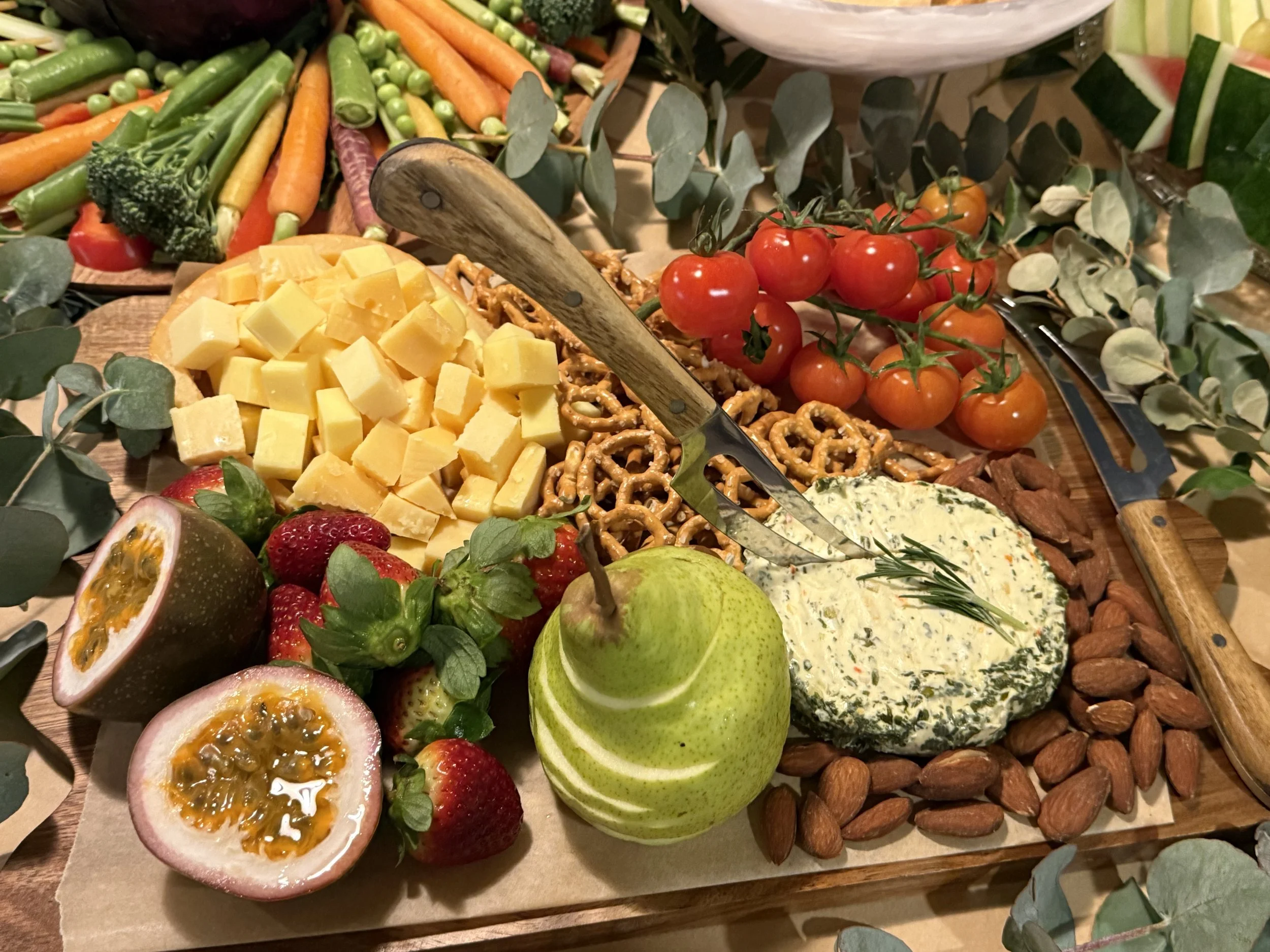 A wooden platter with assorted fresh fruits and vegetables, including cherry tomatoes, cheese cubes, strawberries, passion fruit halves, and a pear, with some leafy greens and crackers, set on a table.