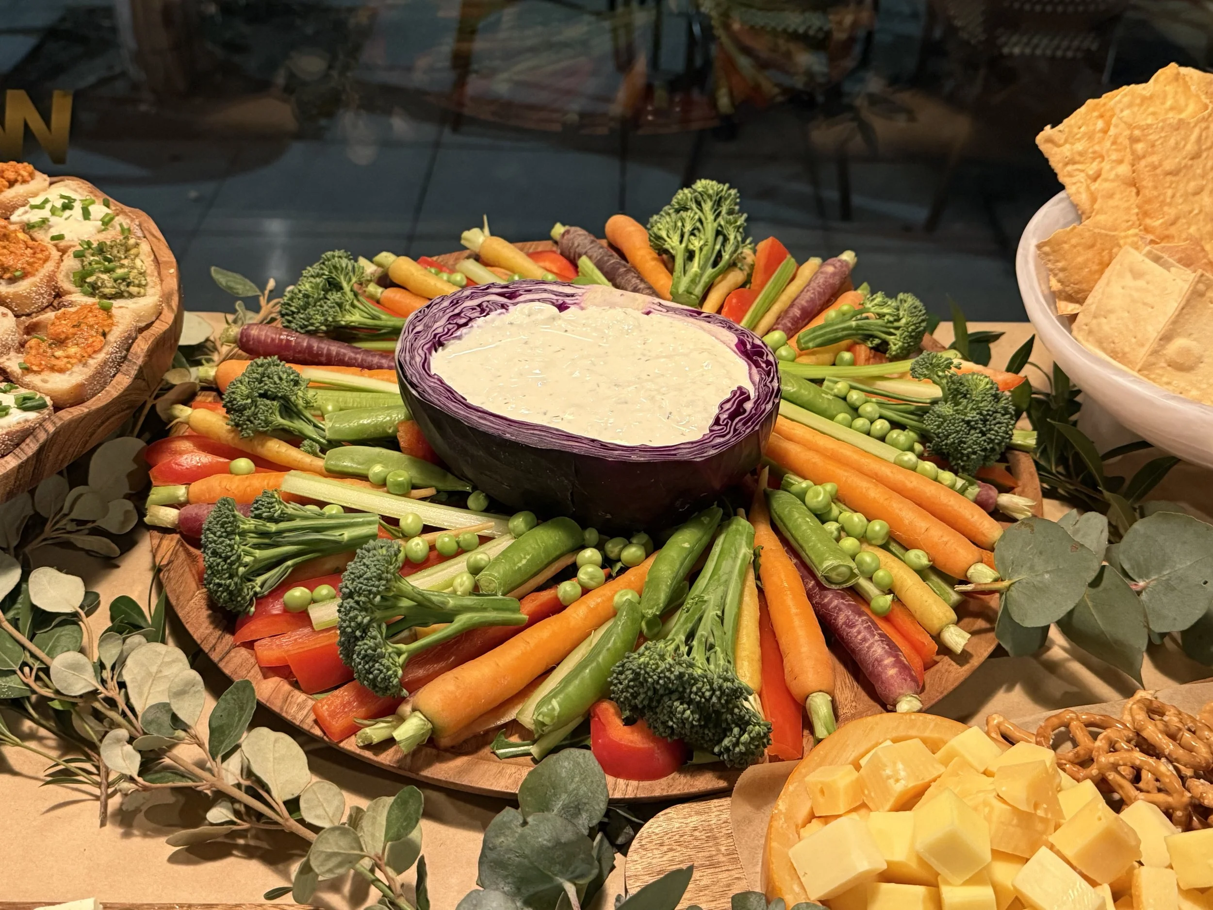 A platter of fresh vegetables including carrots, broccoli, and celery, with a bowl of creamy dip in the center, surrounded by cheese cubes, crackers, and a cheese ball on the side.