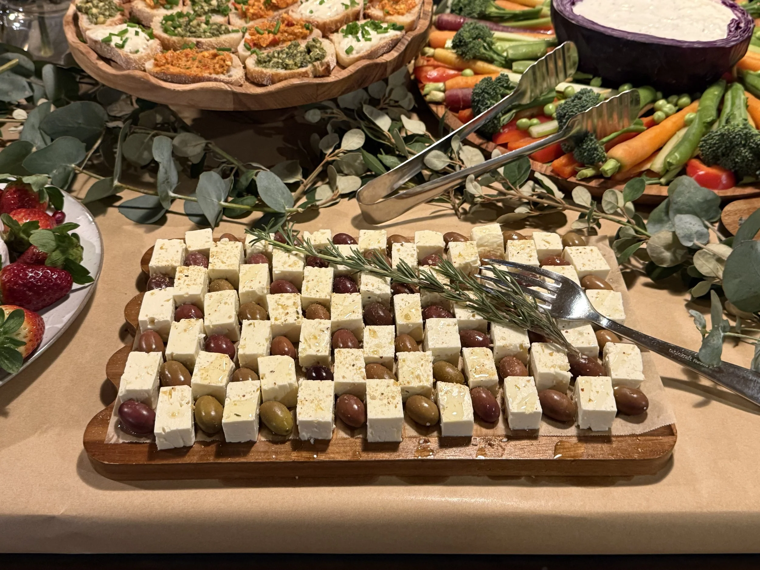 A wooden tray with cubed cheese and green and black olives, garnished with a sprig of rosemary, on a table with other food items including strawberries, vegetables, and dips.