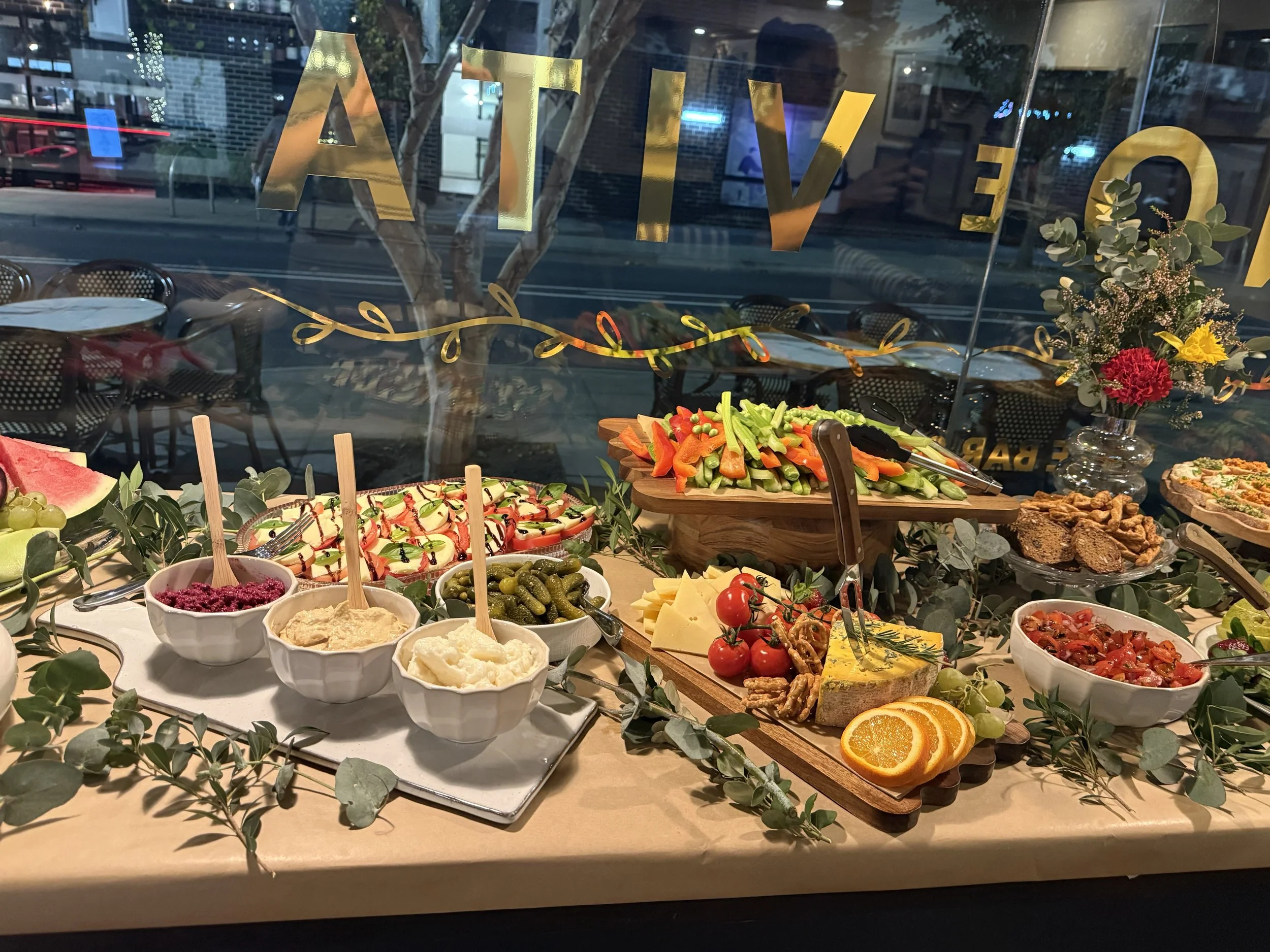 A festive buffet table with a variety of colorful dishes including vegetables, cheeses, fruits, and desserts, decorated with greenery and flowers, situated near a window with city lights outside.