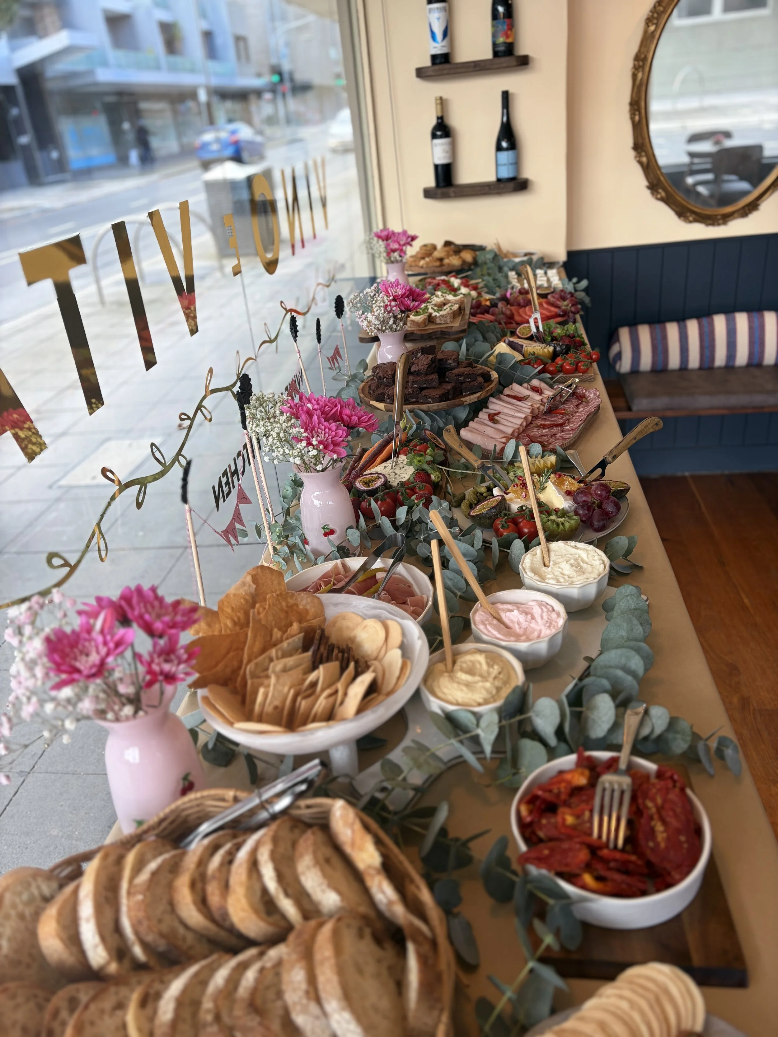 A table with various foods including sliced bread, cookies, dips, fruits, meats, and cheeses, decorated with pink flowers in vases, with a window showing a city street outside.