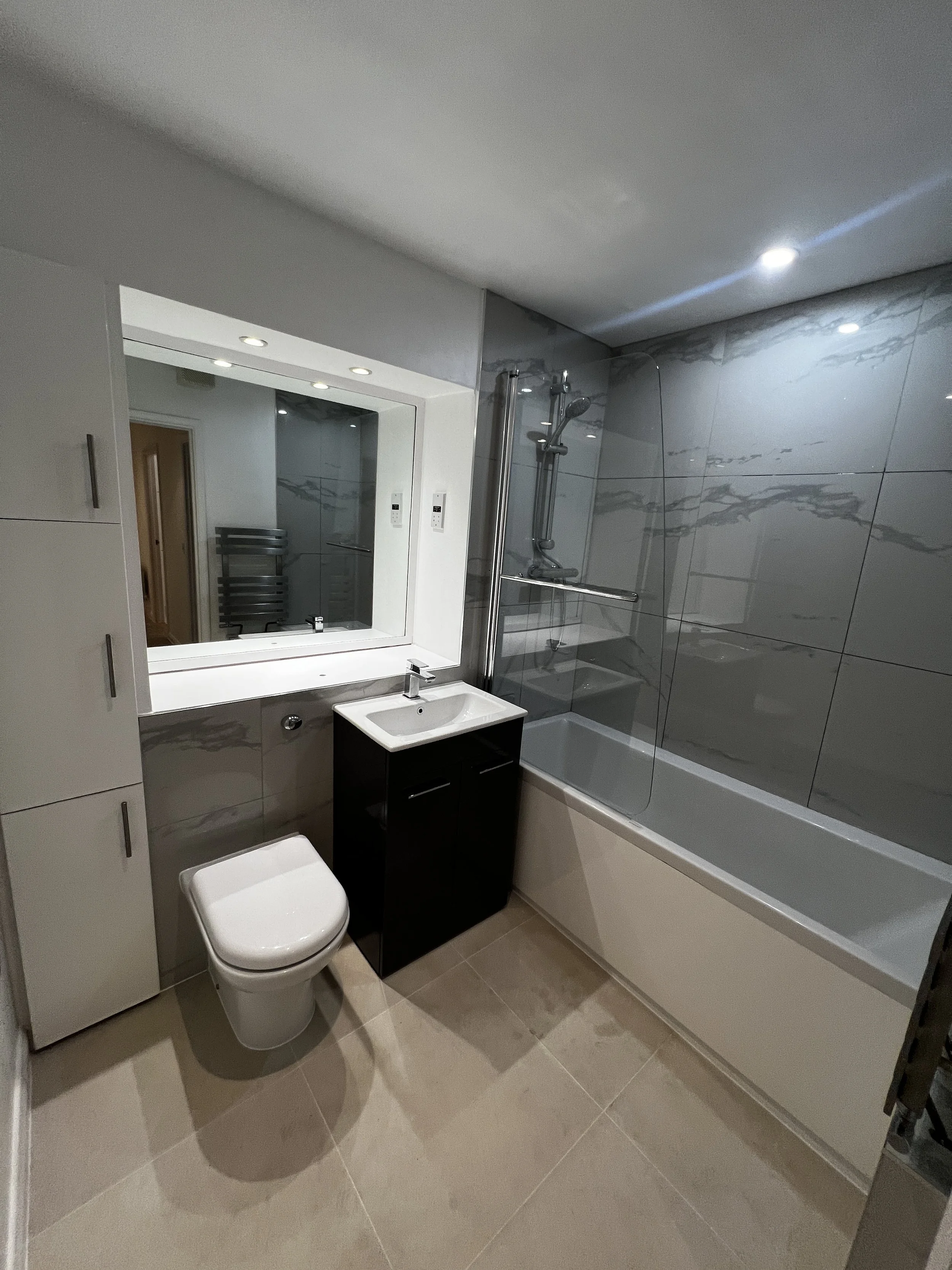 Modern bathroom with a white toilet, black vanity with a sink, large mirror, a bathtub with grey marble tiles, and a showerhead. Recessed ceiling lights illuminate the space.