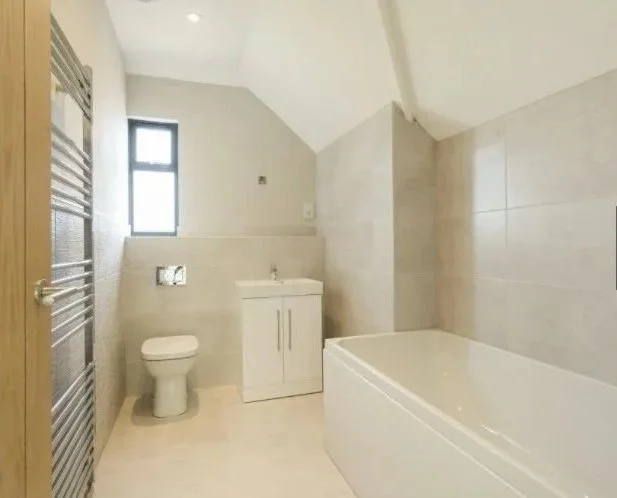 Modern bathroom with a toilet, small sink with cabinet, bathtub, and a window, decorated with light beige tiles.
