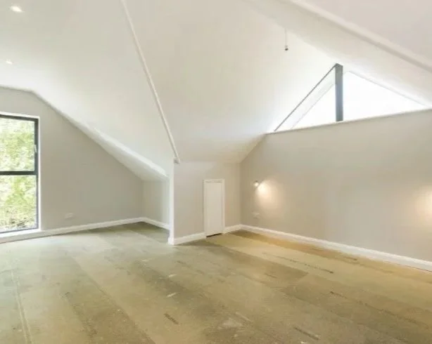 Empty room with sloped ceilings, light gray walls, a window showing greenery outside, and a chandelier.