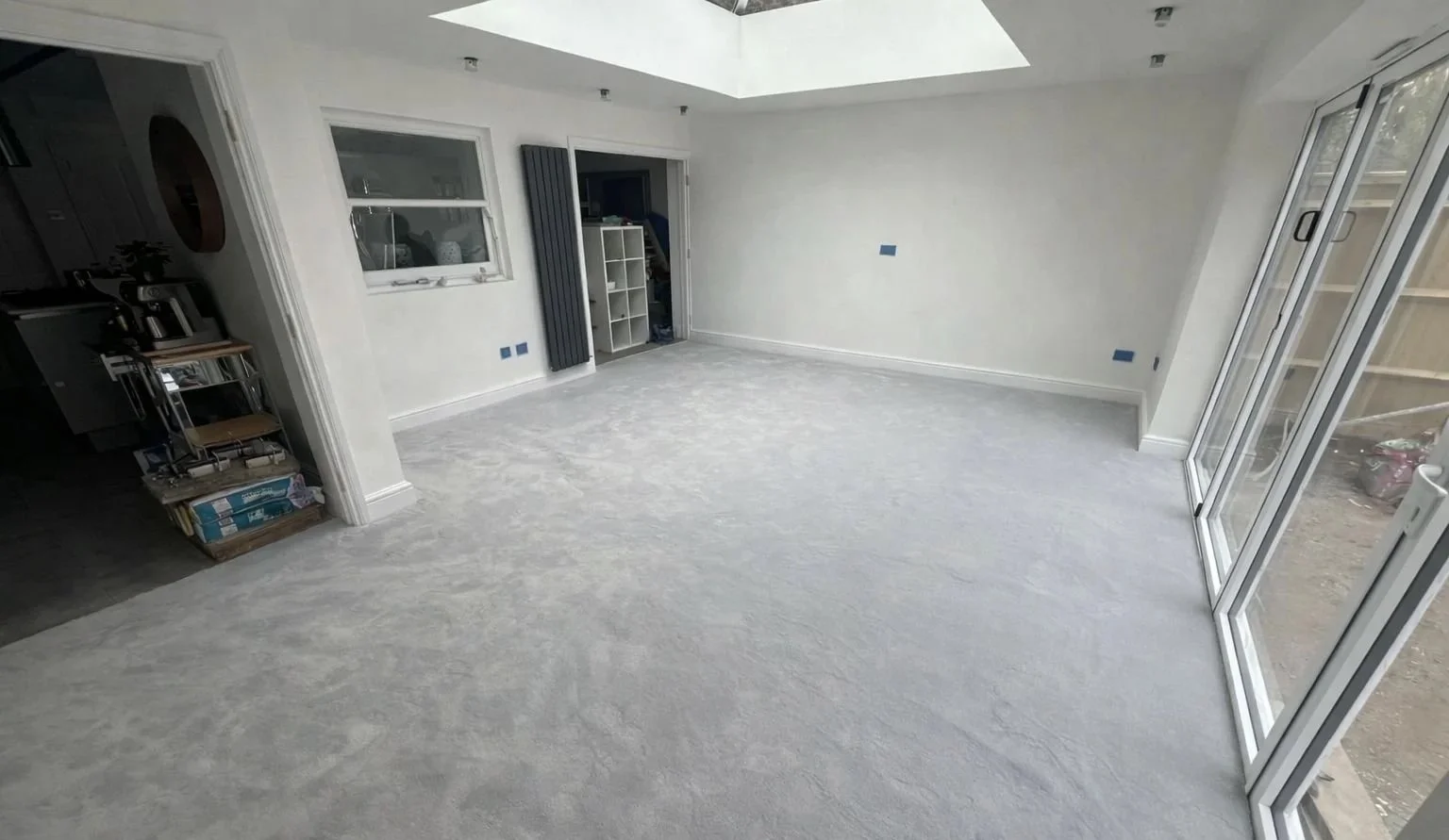 An empty living room with large sliding glass doors on the right, white walls, and a skylight on the ceiling. Light-colored carpeted floor and a small open doorway on the left leading to another room.