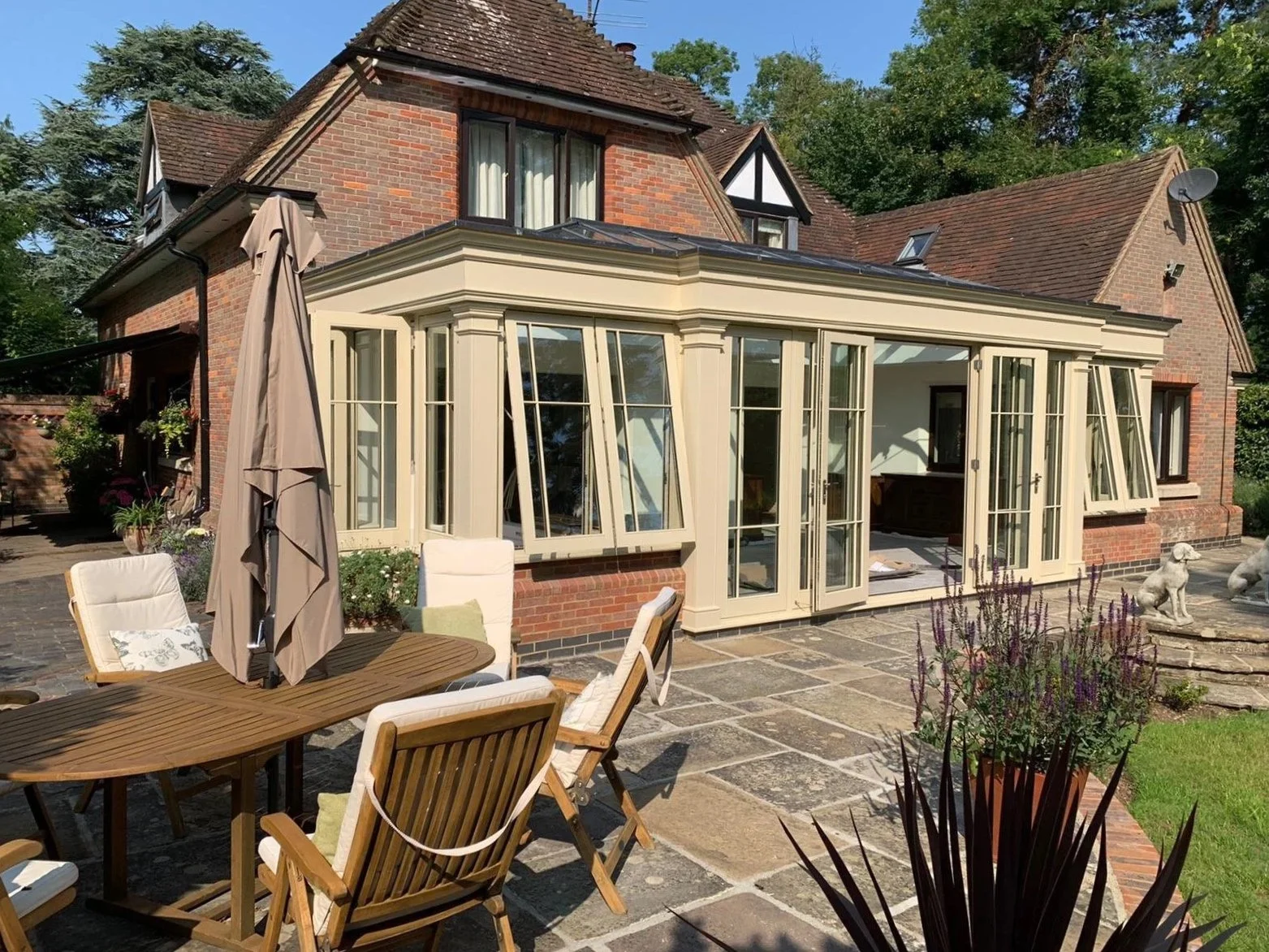 Backyard patio with outdoor dining table, chairs, and large patio doors leading to a house with brick exterior and a sunroom with large windows, surrounded by plants and trees.