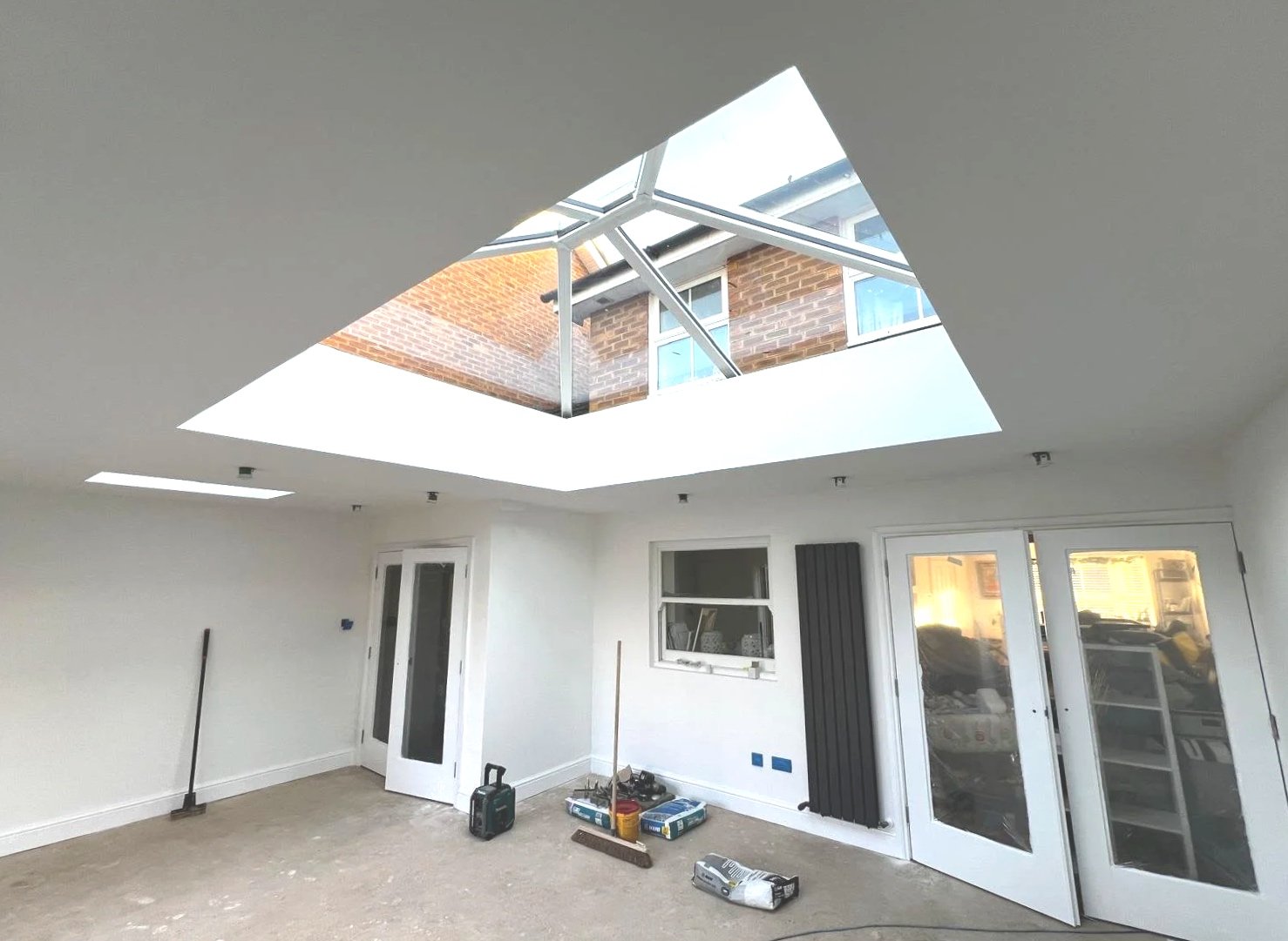 Interior of a room under renovation, featuring a flat ceiling with a large skylight, white walls, and a door with glass panels. The floor is unfinished, with tools and construction materials scattered around.