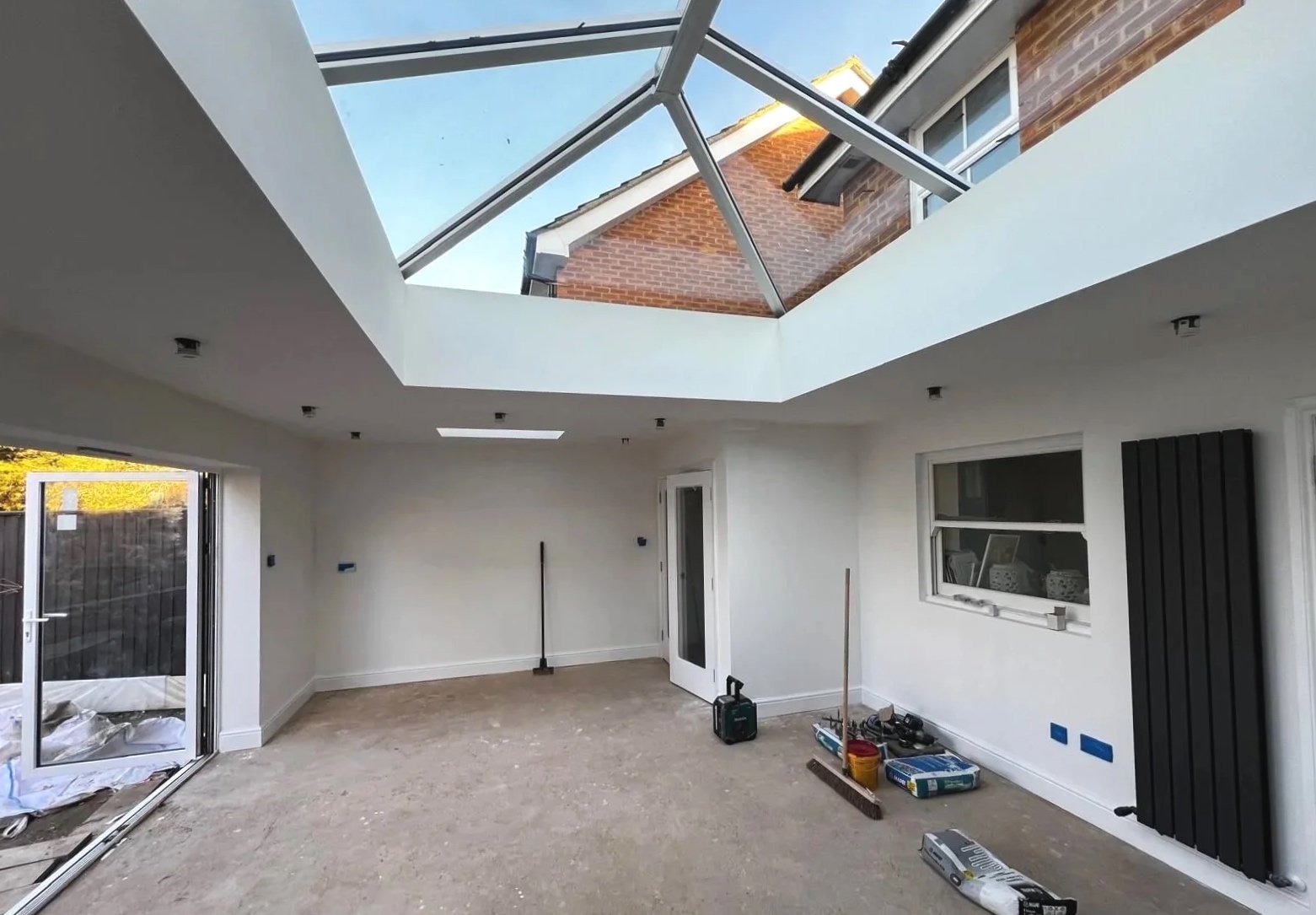 Interior room under construction with a large skylight overhead, white walls, a window, and construction tools and materials on the floor.