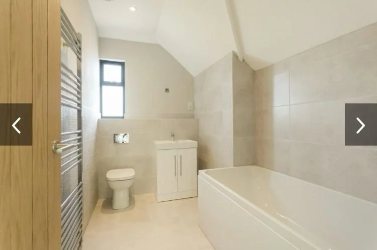 A minimalist bathroom with beige tiles, a white bathtub, a small white vanity with a sink, a toilet, a small window, and a towel heater.