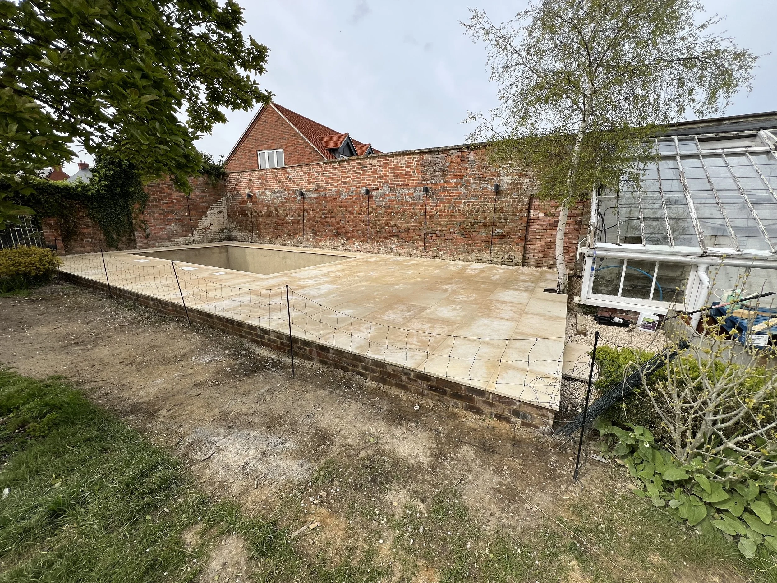 An empty backyard with a partially built or removed swimming pool, a brick wall, a glass greenhouse, trees, and patchy grass.