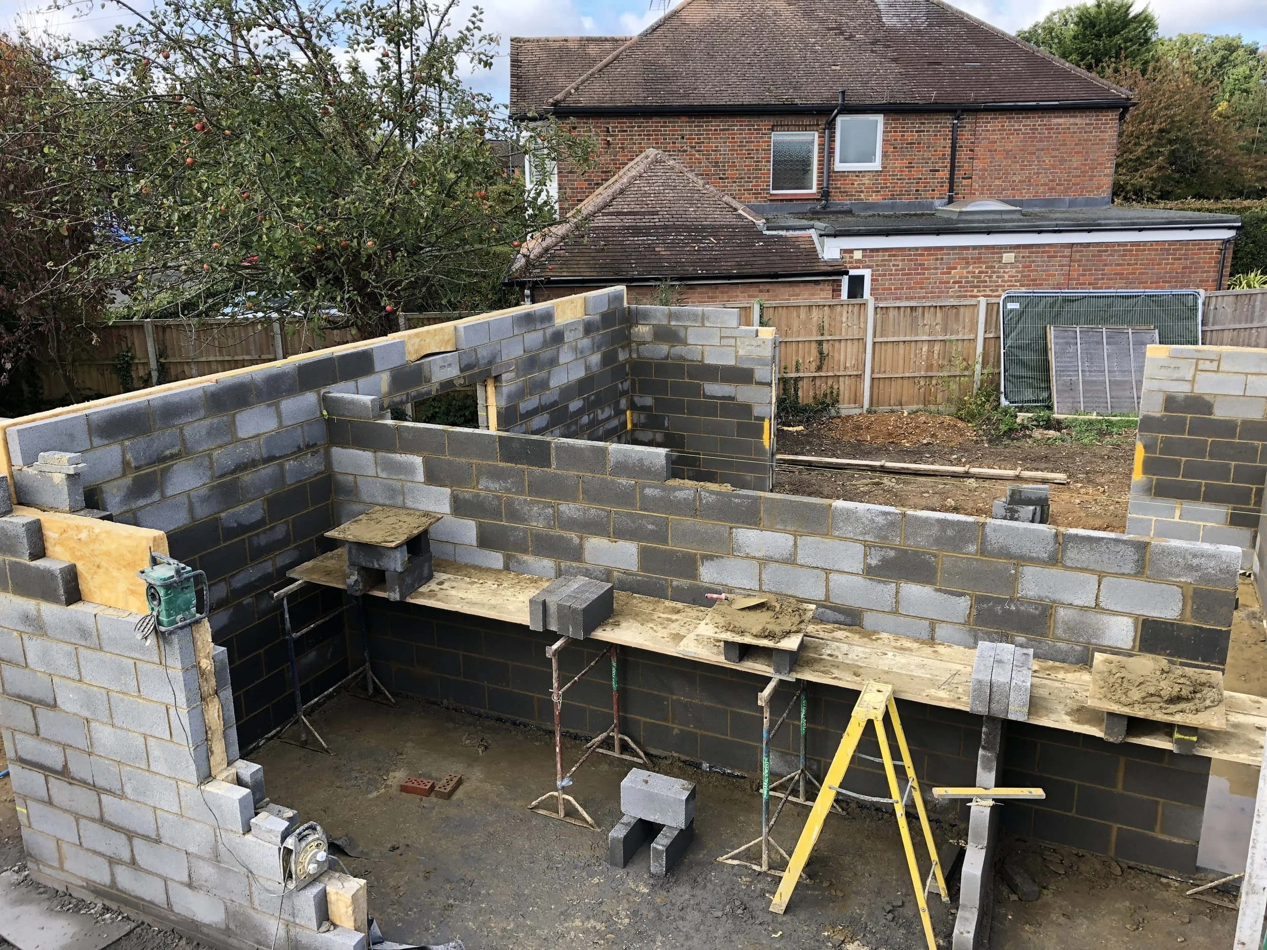 A construction site showing the early stages of building a house, with brick walls partially built and construction tools and scaffolding present.
