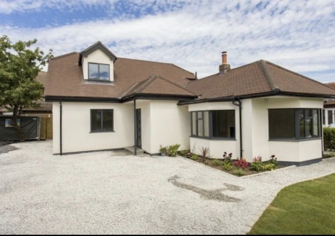 A modern suburban house with white walls, a brown roof, and black window frames, surrounded by a gravel driveway and green lawn.