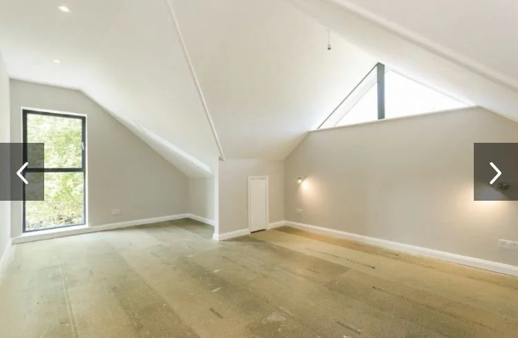 Empty attic room with sloped ceiling, large window, and minimal lighting.