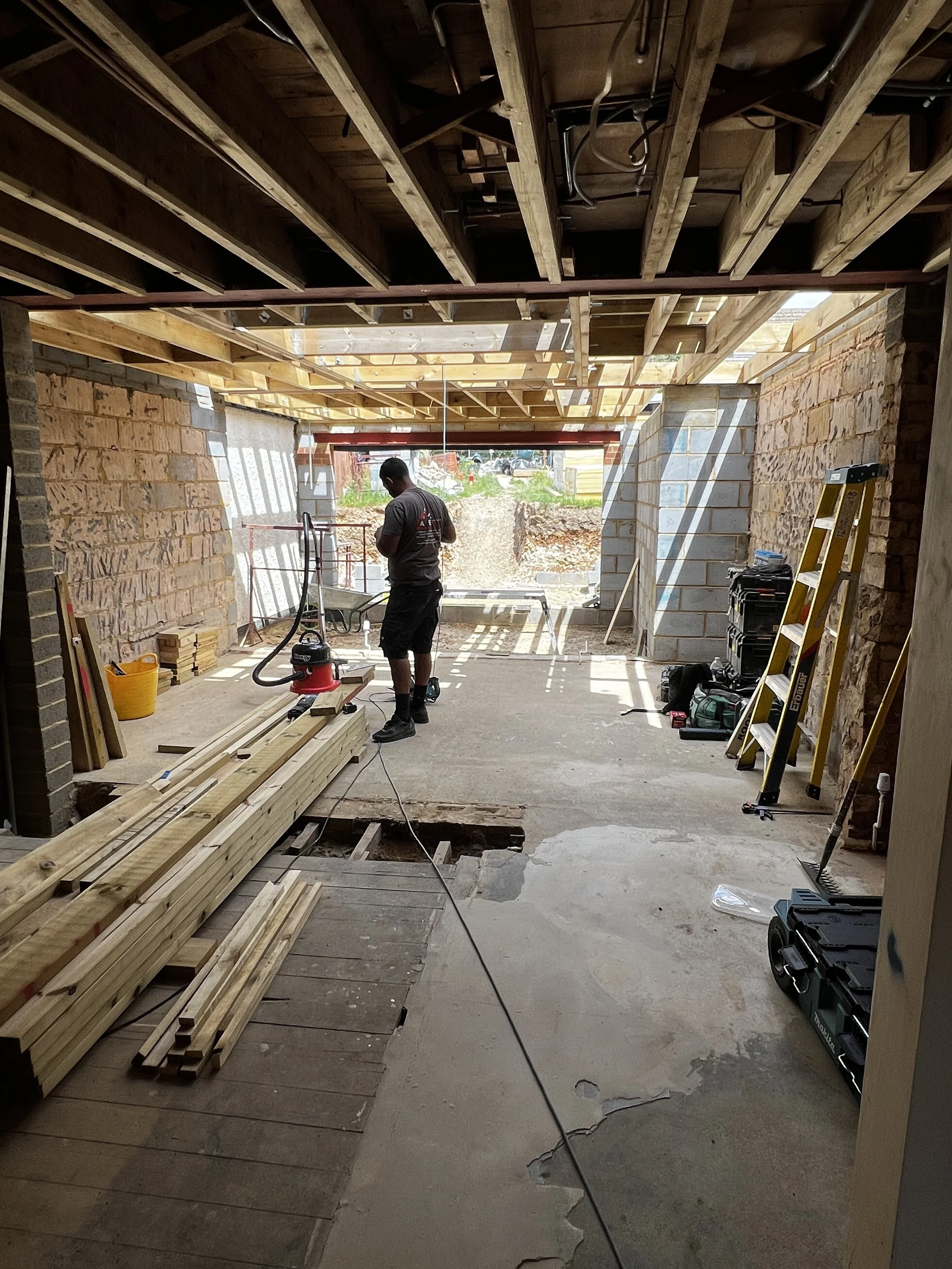Construction worker standing inside a partially built building, with exposed wooden beams and brick walls, surrounded by construction tools and materials.