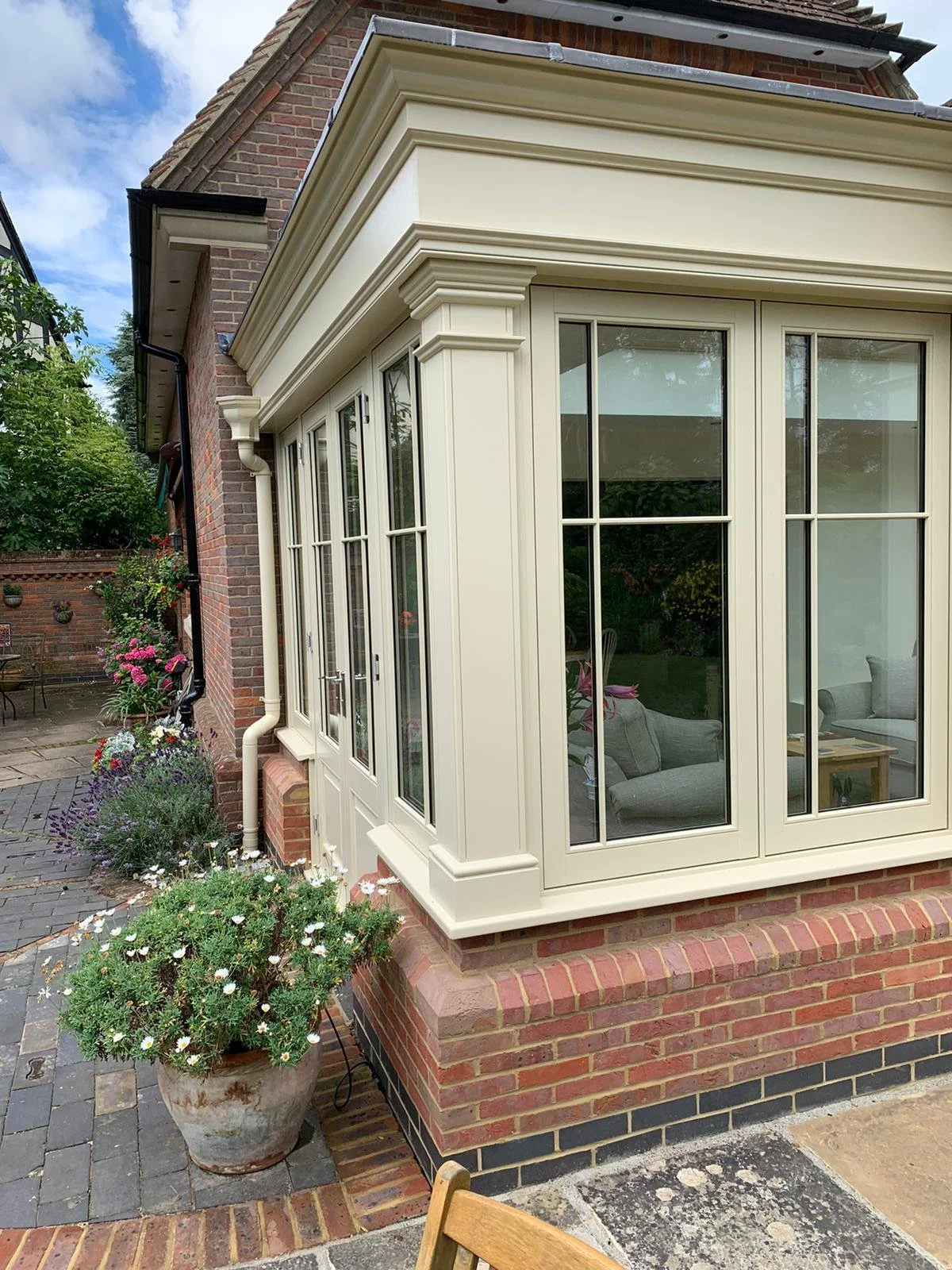 A brick house exterior with a white conservatory featuring large glass windows and doors, surrounded by potted plants and flowers, and garden furniture visible in the background.