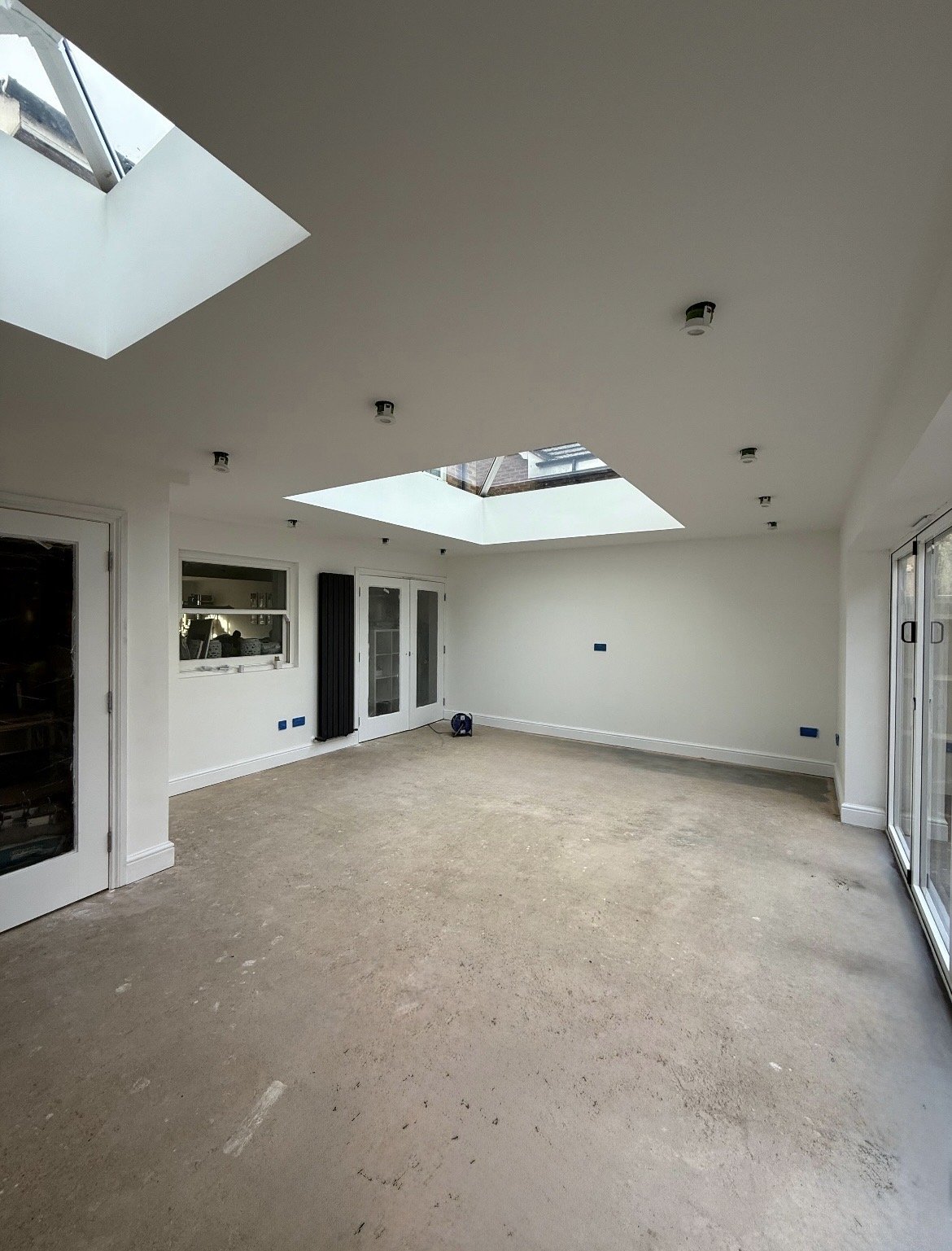 An unfinished modern living room with white walls, large skylights, sliding glass doors, and an exposed concrete floor.