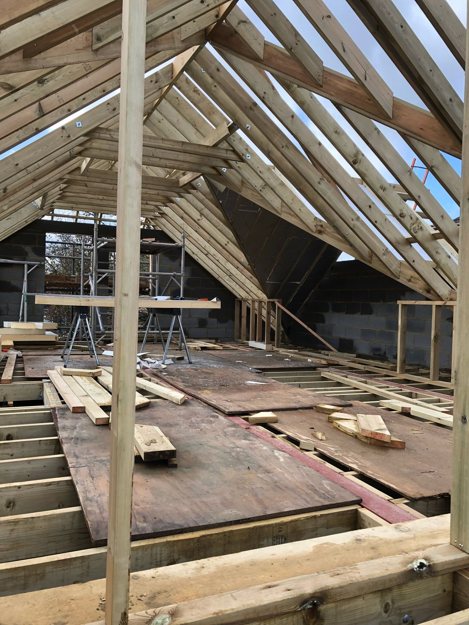 Wooden framing and roof construction of a building under construction, with scaffolding and building materials visible.