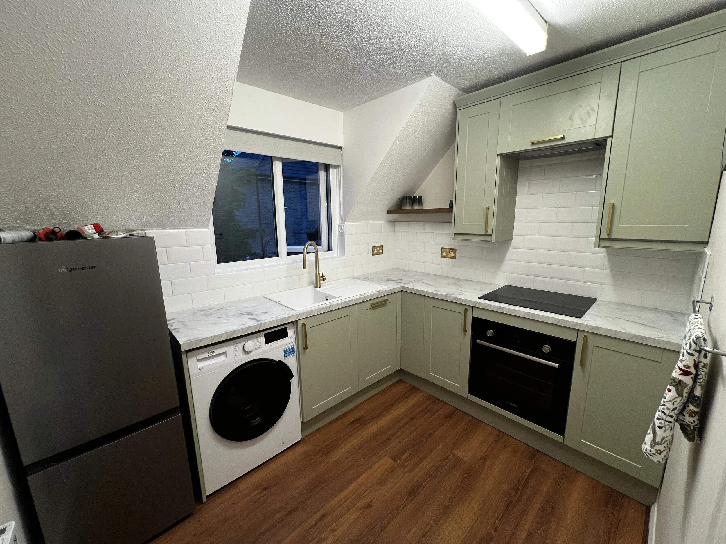 A modern kitchen with light green cabinets, a white marble countertop, white tile backsplash, and a window. There is a black electric stove, a washing machine, and a stainless steel sink with a gold faucet.