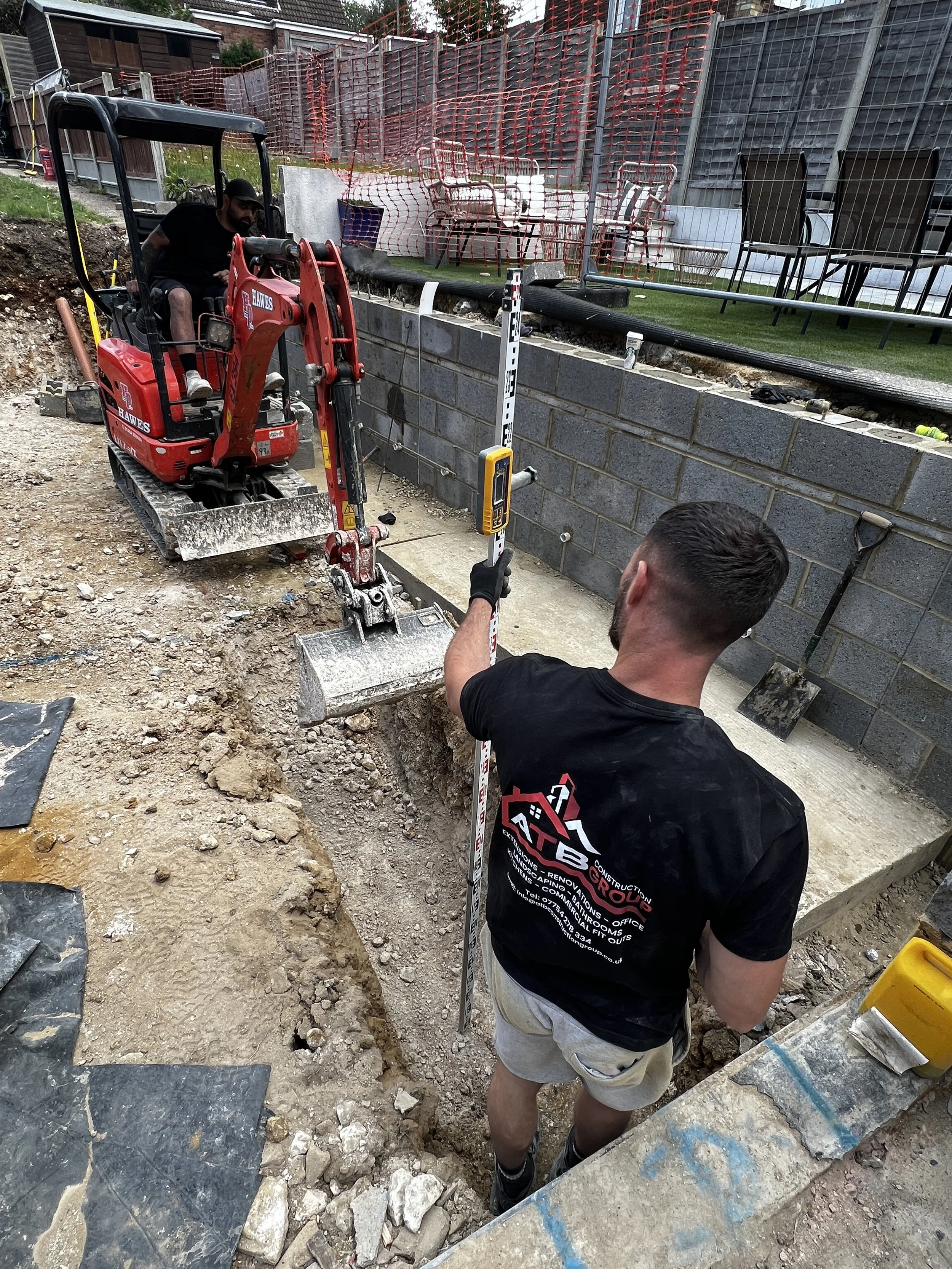 Construction workers laying bricks to build a wall in a backyard, with one man using a spirit level to ensure the wall is straight, and a mini excavator nearby.