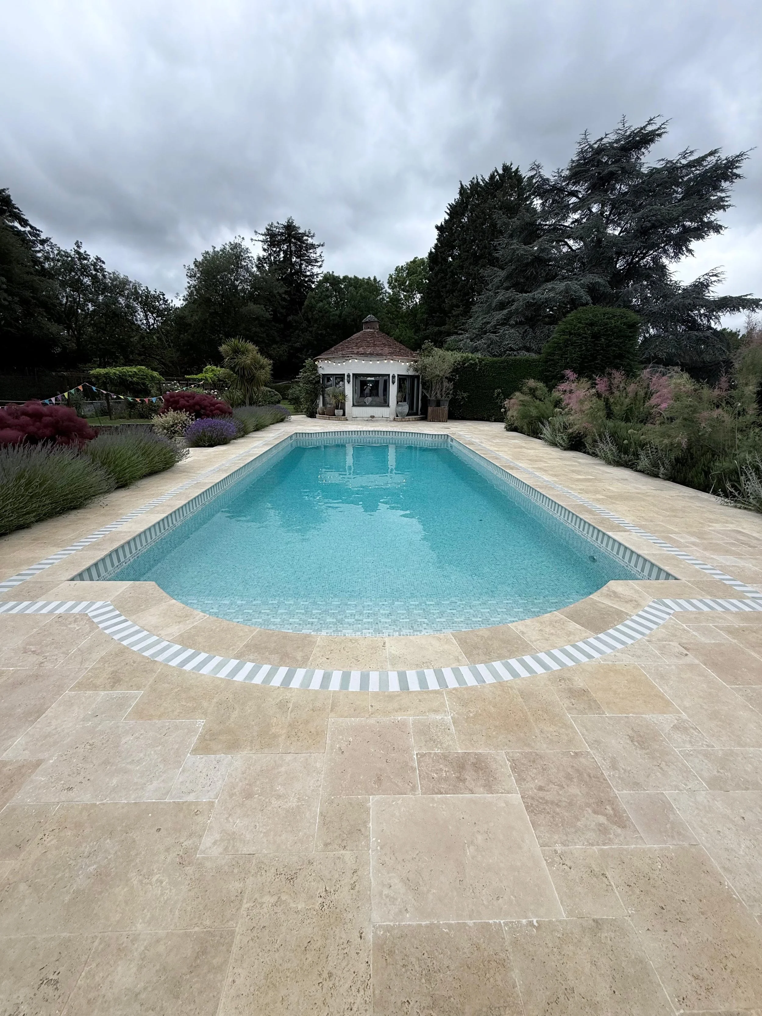 Photo of a backyard with a swimming pool, surrounded by plants and trees, and a small gazebo at the far end under an overcast sky.