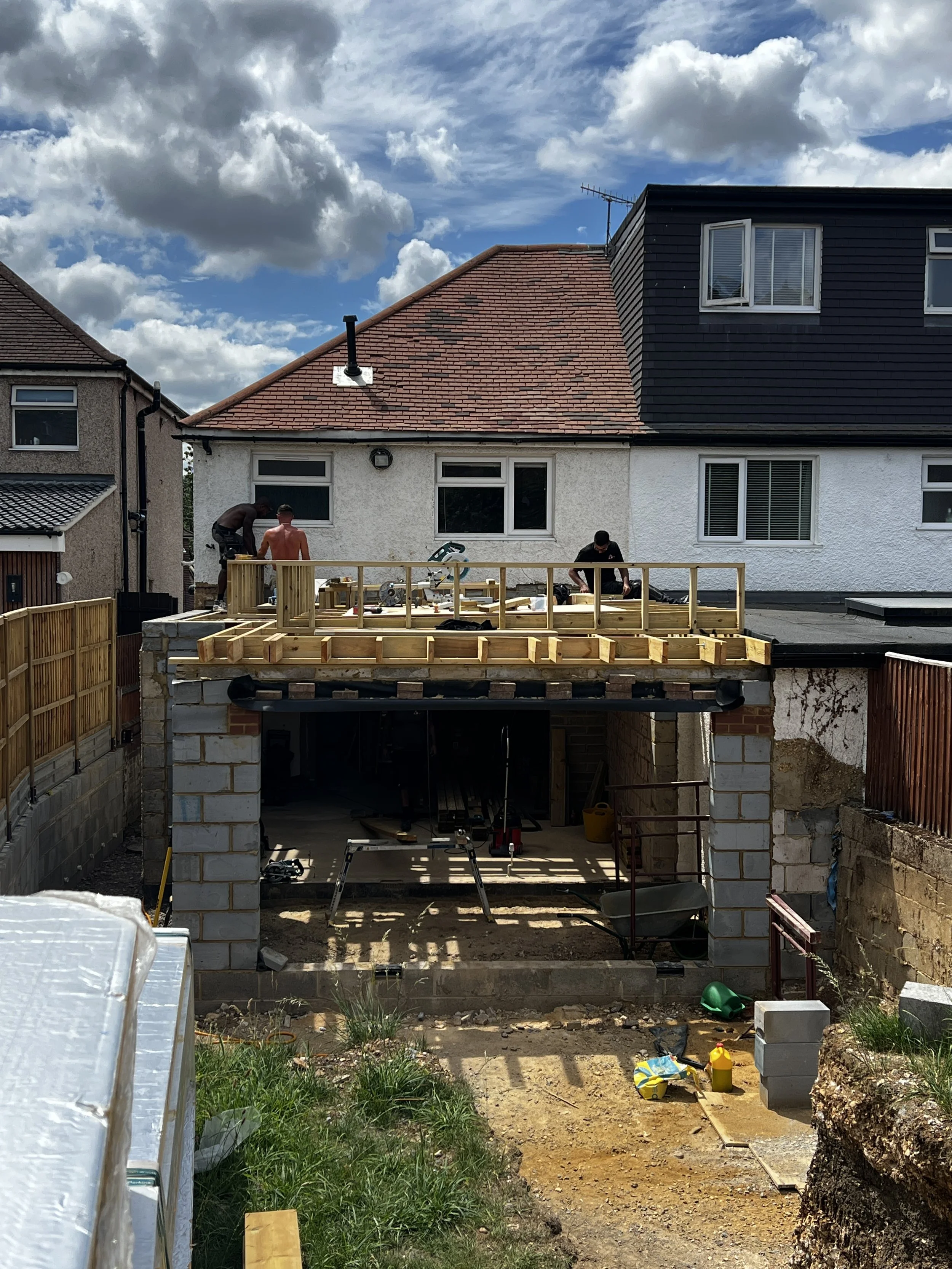 Construction workers building a small extension on a house, with a new wooden platform and some tools around, under a partly cloudy sky.