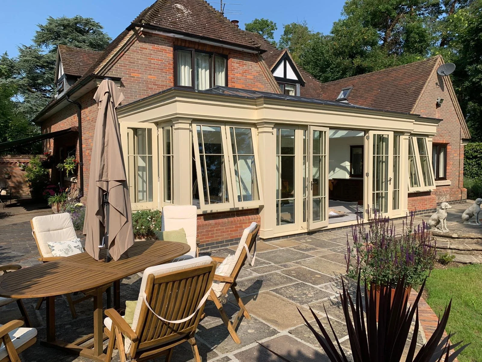 A brick house with a glass conservatory extension in a backyard, with patio furniture, potted plants, and garden statues, under a clear blue sky.