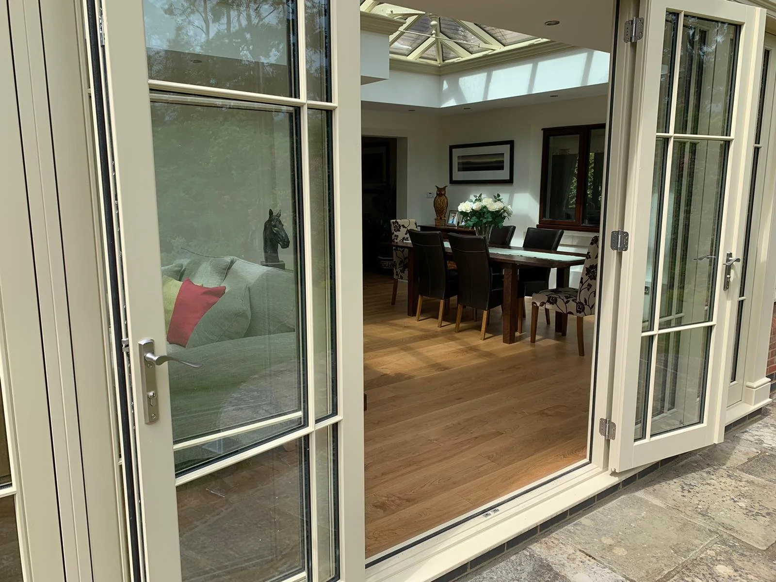 View through open French doors into a dining room with a wooden table surrounded by black and patterned chairs, a vase with white flowers, framed artwork on the wall, and a skylight ceiling.