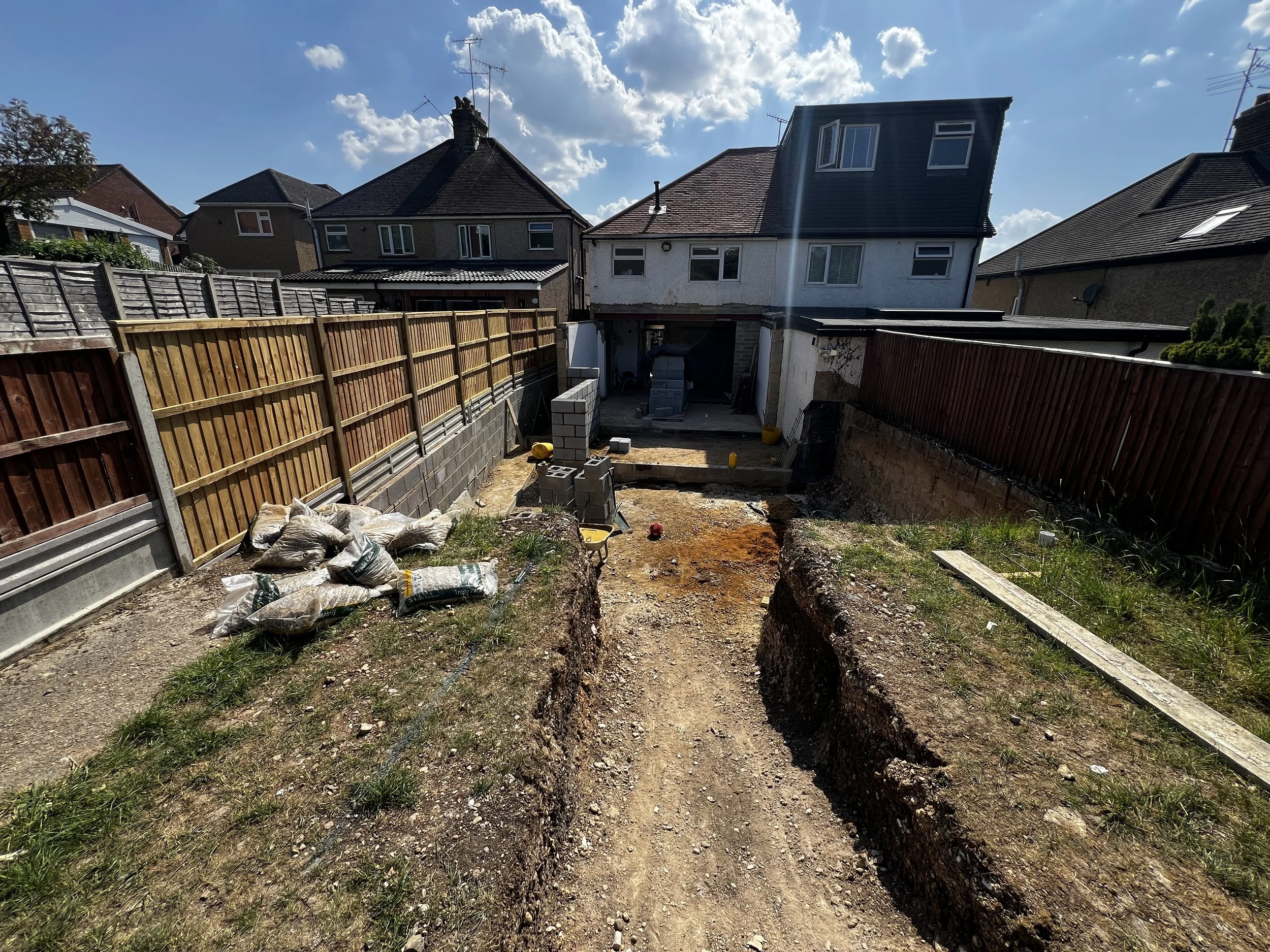 A backyard under construction, showing dirt and trenches with piles of gravel and bricks, surrounded by wooden fences, with neighboring houses and a partly cloudy sky overhead.